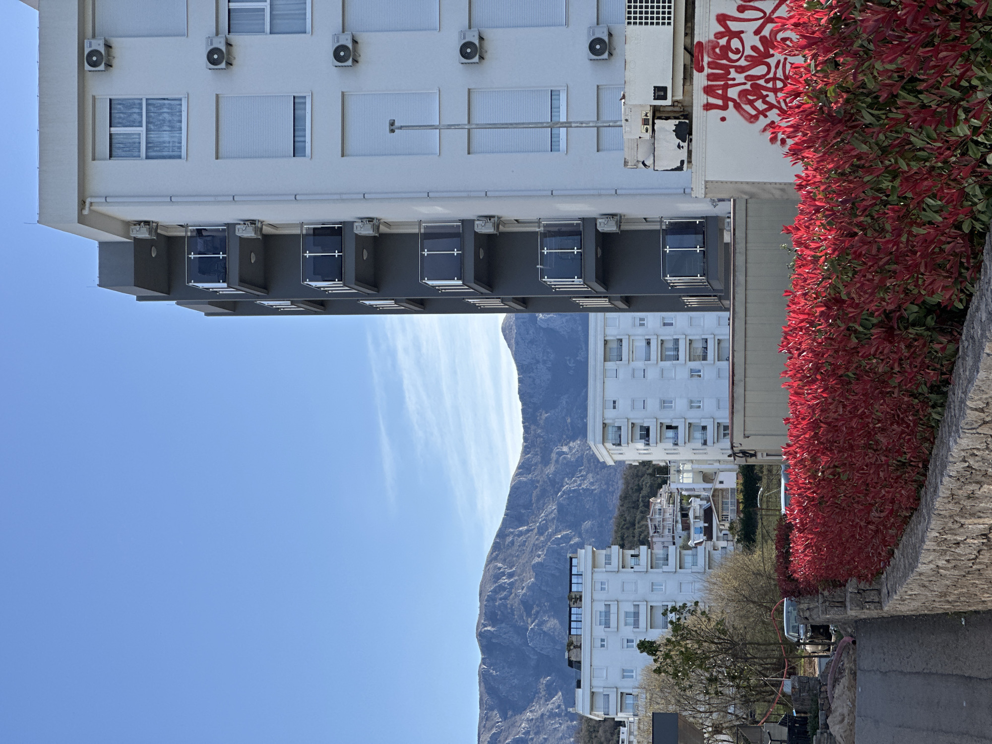 The image presents a scene from Šušanj, Montenegro. A multi-story white building with multiple windows, air conditioning units, and balconies is visible, positioned above the horizon. To the right of the building, a vibrant hedge with red foliage adds a splash of color. In the background, a mountain ridge dominates the skyline. The sky is a clear blue with faint clouds streaking across it. An additional white apartment building and some infrastructure are visible at the base of the mountain. Graffiti is present on the side of one building. A stone wall and part of a road are in the foreground.