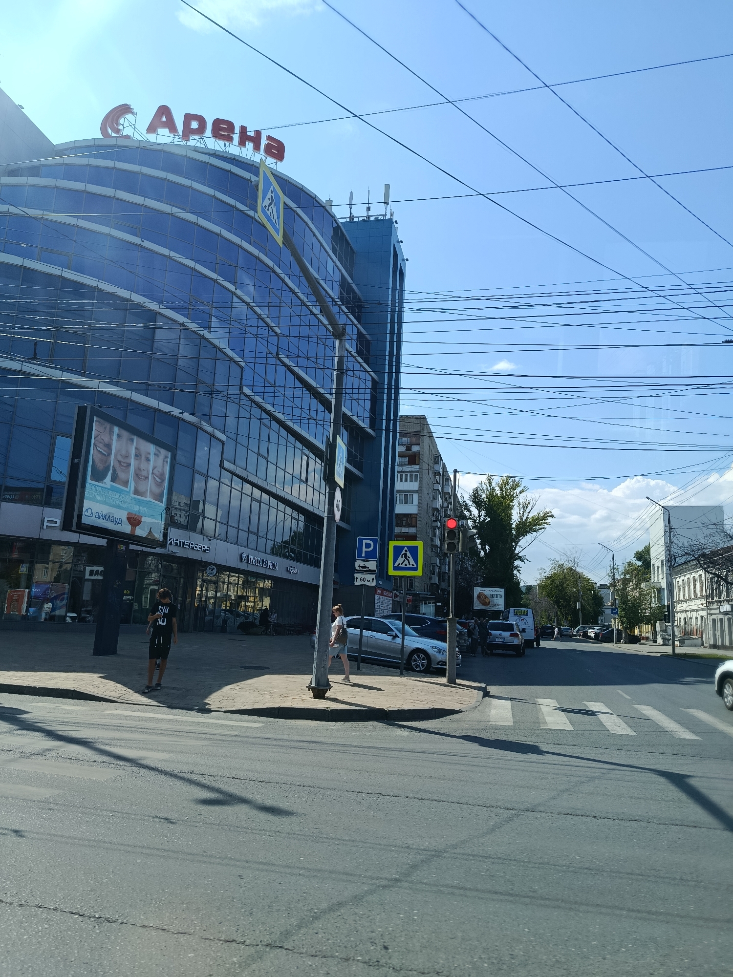 The image depicts a street scene in Saratov, Russia, likely during daylight hours under a clear blue sky with scattered clouds. The foreground is dominated by an asphalt road with a pedestrian crossing. A light pole with traffic signals and a pedestrian crossing sign is positioned on the right side of the road.
To the left of the road is a large, modern building with a curved facade predominantly covered in blue glass. A prominent red sign at the top of this building reads "Арена" (Arena). In front of the building, a digital billboard displays images of smiling faces and text. Sidewalks are visible along the building, with a few individuals walking or standing. One person, a young male in dark clothing, is walking on the sidewalk, and another, a young female in light clothing, is also on the sidewalk. Several parked cars are visible along the street and in front of the building.
Further down the street, other buildings are visible, including a multi-story residential building with balconies and an older, white-facade building with decorative architectural elements on the right. Overhead, a complex network of power lines crisscrosses the sky. The overall impression is a bustling urban environment with a mix of modern and older architecture.