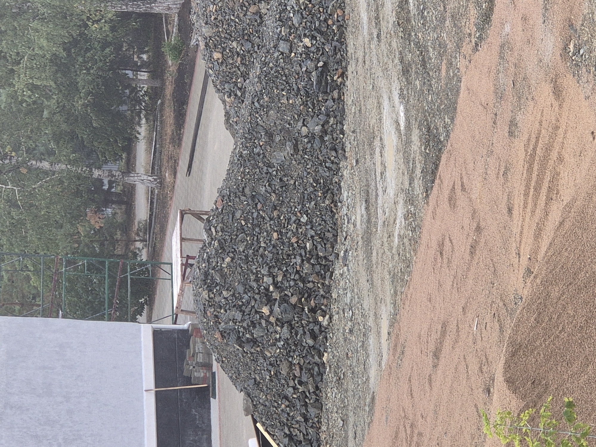 This is a medium-shot, outdoor photograph taken on an overcast day. The foreground features a large, irregularly shaped pile of dark grey gravel. To the left and slightly behind the gravel pile, there is a stack of bricks. A wooden broom leans against a light-colored wall, which is partially visible on the far left of the image. Behind the gravel pile and bricks, a section of scaffolding with green metal supports is visible, situated next to the light-colored wall. Beyond the scaffolding, a paved pathway or road is visible, lined with trees and foliage. In the mid-ground, another pile of gravel can be seen. The background consists of trees and the overcast sky. The scene appears to be a construction or landscaping site. The location is Kokshetau, Kazakhstan.