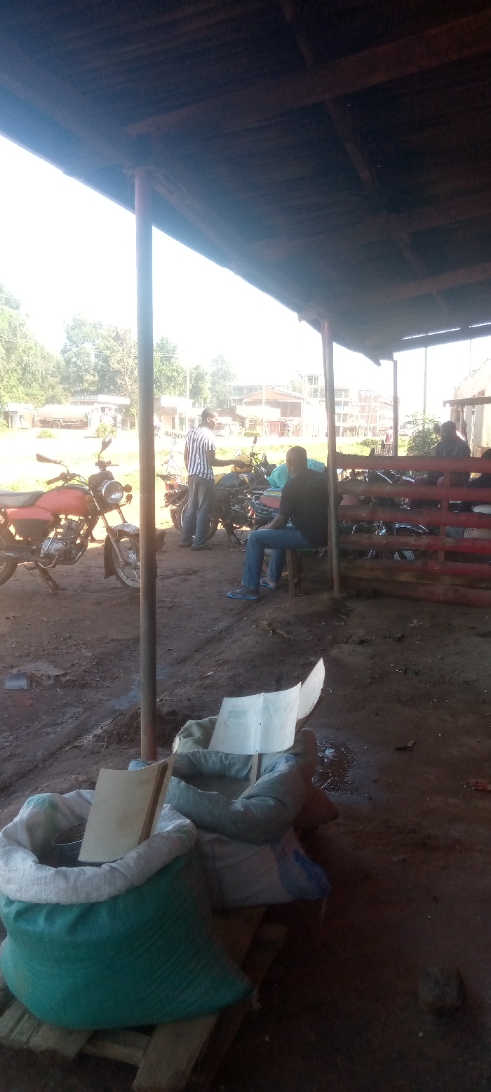 The scene is taken under a corrugated iron roof in a village in Kivaywa, Kenya. The ground is dusty. There is a man in a black and white shirt, he appears to be interacting with a man sitting on a chair. The man sitting on the chair is wearing a blue shirt, jeans and sandals. There are numerous motorbikes in the scene. There is a large green bag with what looks like a white book on top of it, and a white bag next to it. There is a small piece of paper on the ground. The image was likely taken from a slightly elevated angle. The location is close to the coordinates 0.56, 34.806. The scene shows a typical gathering place for men in a rural Kenyan community.