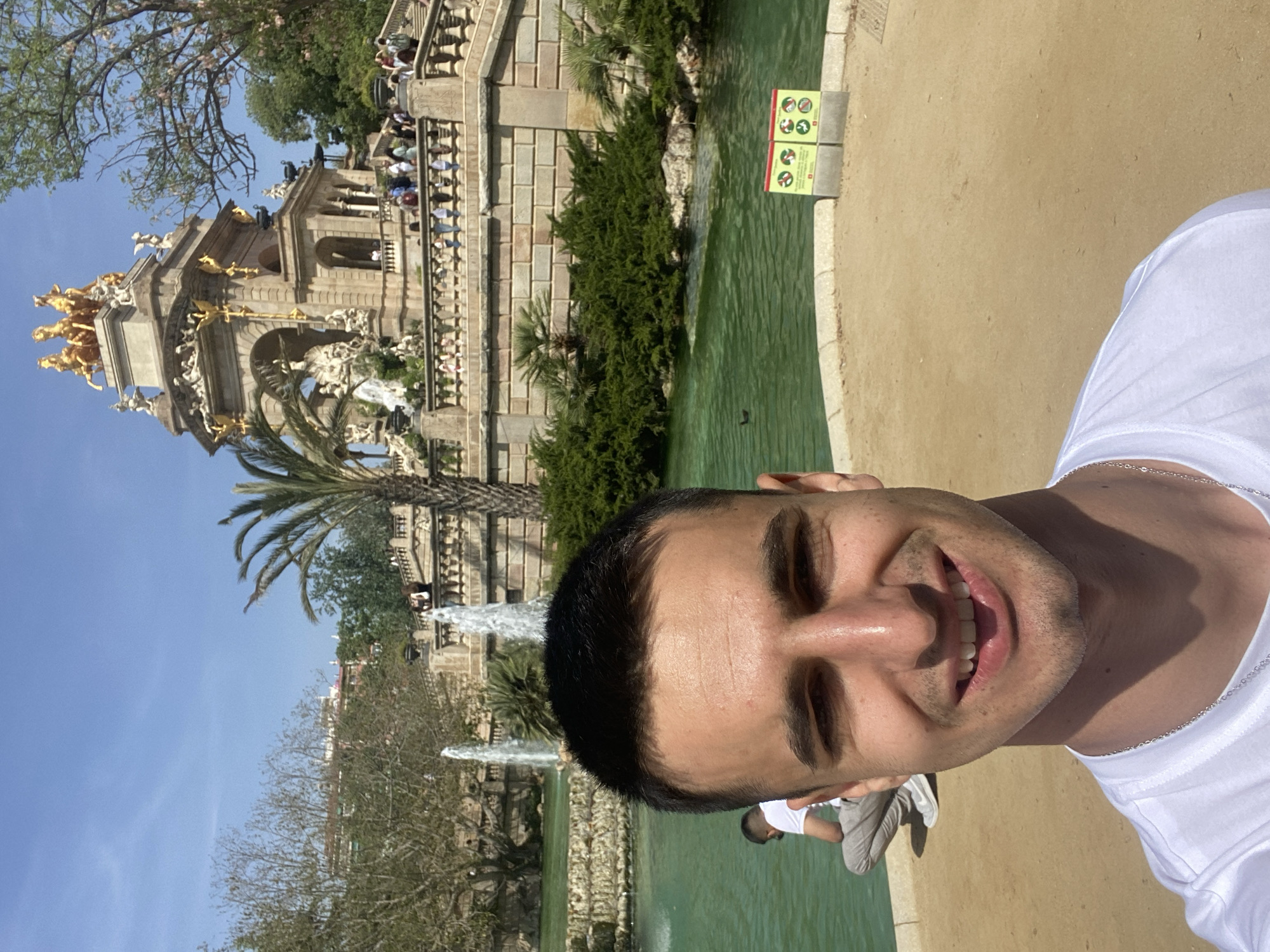 The image is a bright, sunny selfie taken by a smiling man in an outdoor park setting, identified as Barcelona, Spain. The man, visible from the chest up on the right side of the frame, has short dark hair and is wearing a white t-shirt and a silver chain around his neck. He appears happy, looking directly at the camera.
In the background, dominating the scene on the left, is the grand Cascada Monumental (Monumental Waterfall) in Parc de la Ciutadella. This ornate fountain features multiple levels of white stone architecture and intricate sculptures, highlighted by prominent golden statues, including a large quadriga (chariot pulled by four horses) at its highest point. Water can be seen cascading down various sections of the monument. Many people are visible on the steps and terraces of the fountain, suggesting it is a popular tourist attraction.
In the mid-ground, a body of green-tinged water, likely a pond or canal, extends across the scene. The water's edge is lined with green shrubs and trees, including a tall palm tree near the fountain. A sandy or gravel path runs alongside the water on the right. The sky is a clear, bright blue, indicating sunny weather, and strong shadows suggest it is midday or early afternoon.
A yellow sign with multiple red-circled prohibition symbols is visible near the water's edge on the right. These symbols indicate restrictions such as "no swimming," "no feeding animals," and "no littering." Another individual, partially visible in the lower right background, is seated or crouching near the water.