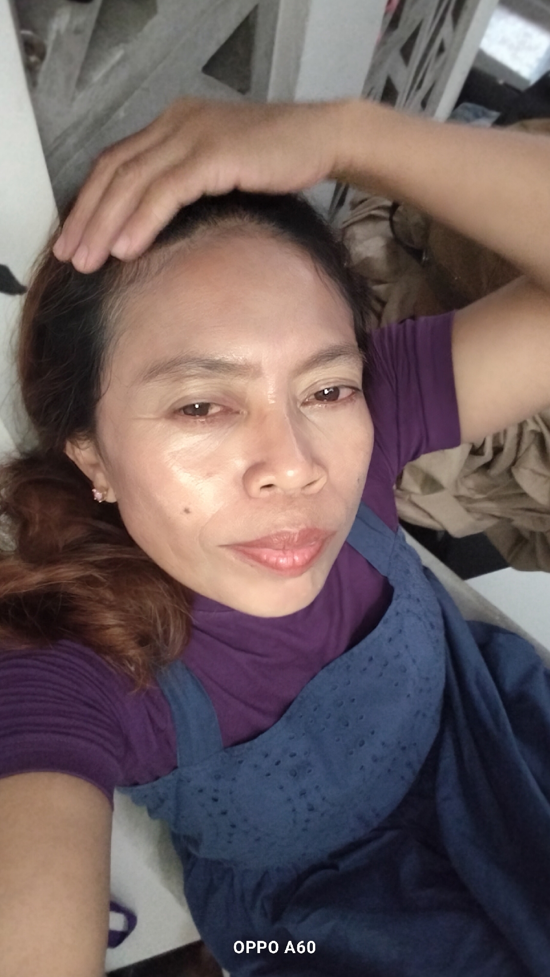 The image is a close-up selfie of a woman in what appears to be an indoor setting, possibly a bedroom or living room in North Kuta, Indonesia. The woman, who has brown hair and is wearing a purple top and a blue pinafore dress, is lying down and has her arm raised, touching her head. Her face is the primary focus, with her eyes open and looking towards the camera. The lighting suggests it is daytime. In the background, a patterned surface and some folded fabric are visible, indicating the presence of furniture or bedding. The bottom of the image displays "OPPO A60" in white text, suggesting the photo was taken with an OPPO A60 mobile phone.