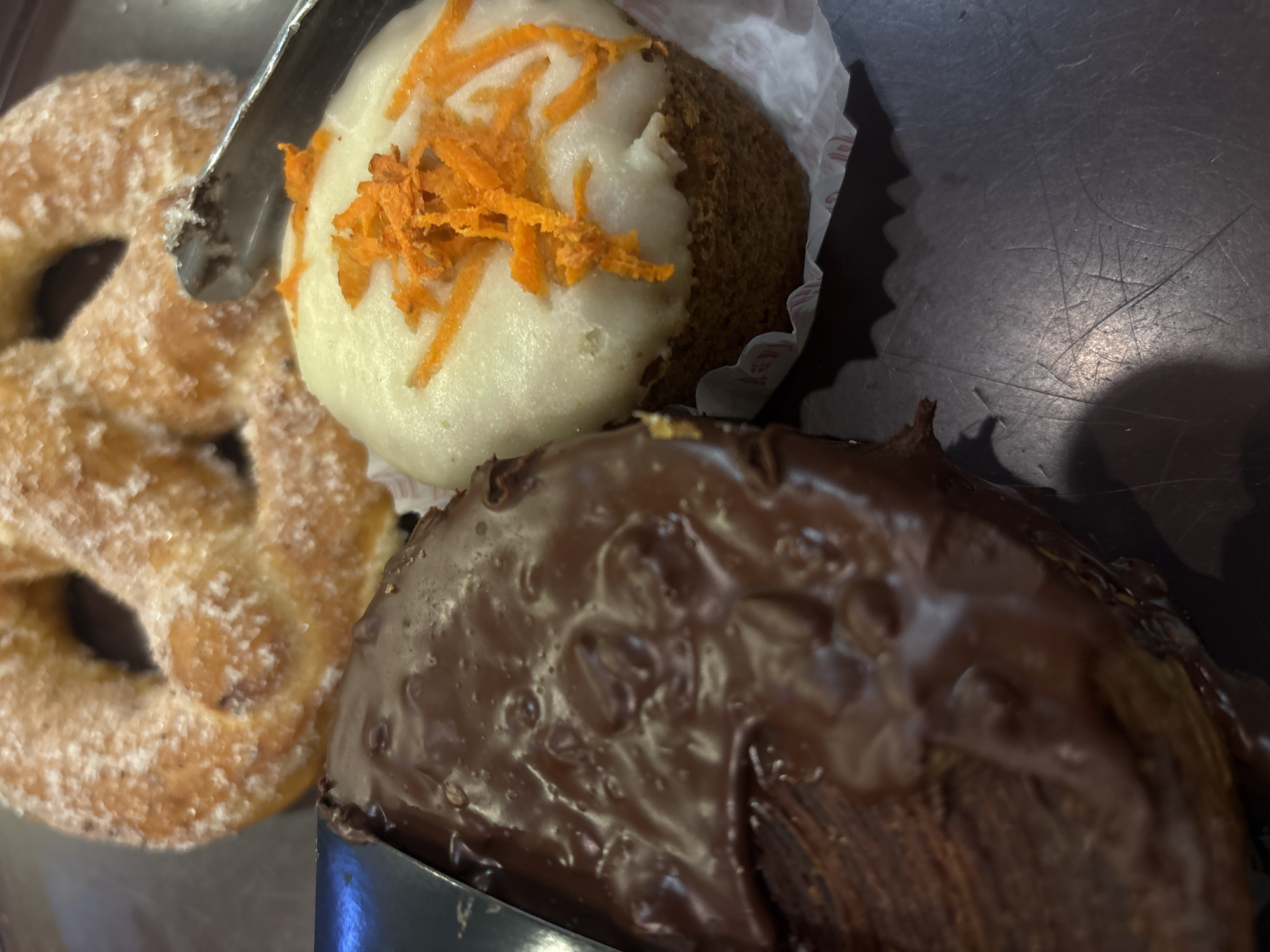 The image shows a close-up of three pastries on a dark surface. From left to right, there is a sugar-dusted pretzel, a carrot cake cupcake with white frosting and shredded carrot topping, and a chocolate-glazed pastry, likely a croissant or similar viennoiserie. A pair of metal tongs is partially visible, positioned near the pretzel and cupcake. The pastries appear to be from a bakery in Mexico City, Mexico.
