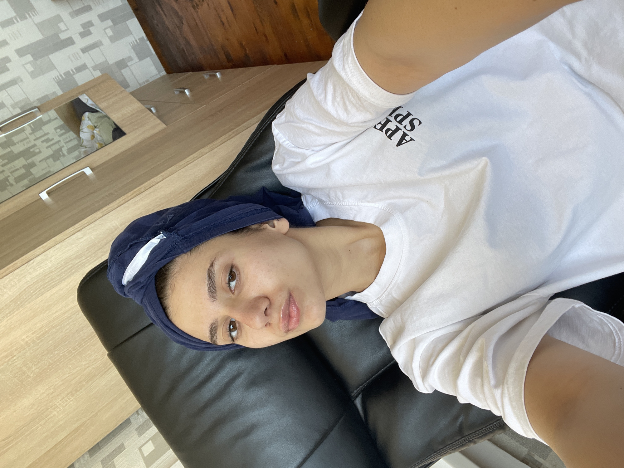 A young woman is lying on a black leather sofa. She is wearing a white t-shirt and a dark blue head covering with a white stripe. The background includes a wooden cabinet or wardrobe with silver handles and a patterned wallpaper in shades of grey and white. A portion of the floor is visible, showing light-colored tiles. The photo appears to be taken indoors, possibly in a residential setting. The location cannot be determined to be in Sofia, Bulgaria, from the provided image.