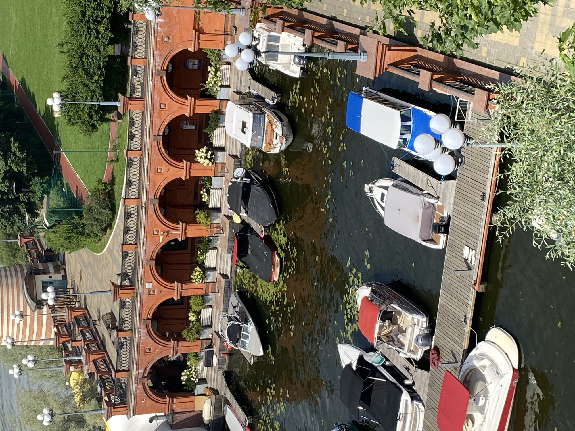 An aerial view captures a marina with several boats docked along a wooden pier. The boats vary in size and design, with some featuring canopies and covers. The water is calm and dotted with lily pads. In the background, a red brick building with arched walkways is visible, adorned with potted plants. A paved walkway with decorative railings and streetlights runs alongside the building. Beyond the building, a lush green lawn and a red running track are present. The scene appears to be a well-maintained waterfront area with recreational facilities.