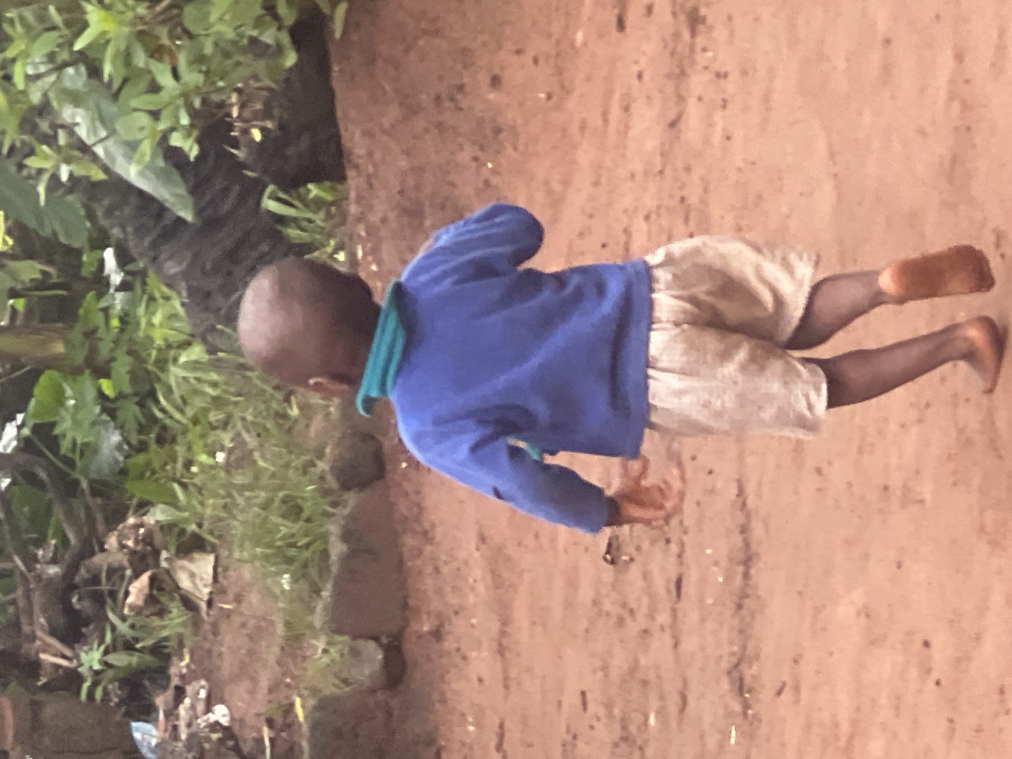 A young child, viewed from behind, runs across a dusty, reddish-brown dirt path. The child is wearing a blue long-sleeved shirt with a teal collar and light-colored, loosely fitting shorts. Barefoot, the child's legs are visible, with the right foot appearing to be mid-stride, slightly raised. The background is a blur of green foliage and dark, tree-like shapes, suggesting an outdoor, possibly rural or semi-urban environment. In the upper left corner, some debris including what appears to be a plastic bag is visible. The scene is likely located in Onitsha, Nigeria, given the context provided.