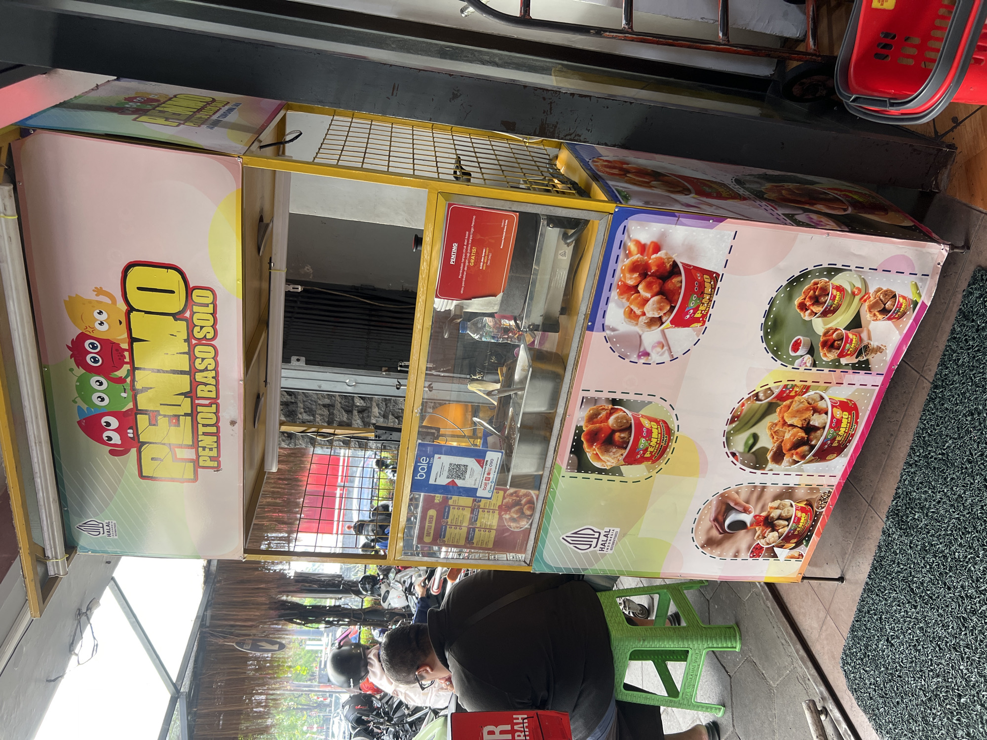 The image shows a food stall in Yogyakarta, Indonesia, selling "Pentol Baso Solo." The stall is adorned with colorful graphics featuring cartoon characters and images of the food product. A person is seated on a green plastic stool in front of the stall, looking away from the camera. The stall itself is a portable structure with a yellow metal frame and a transparent display case containing food items. A menu and a QR code are visible on the display case. The background shows a street scene with parked motorcycles and buildings. The overall impression is of a bustling street food vendor in a Southeast Asian city.