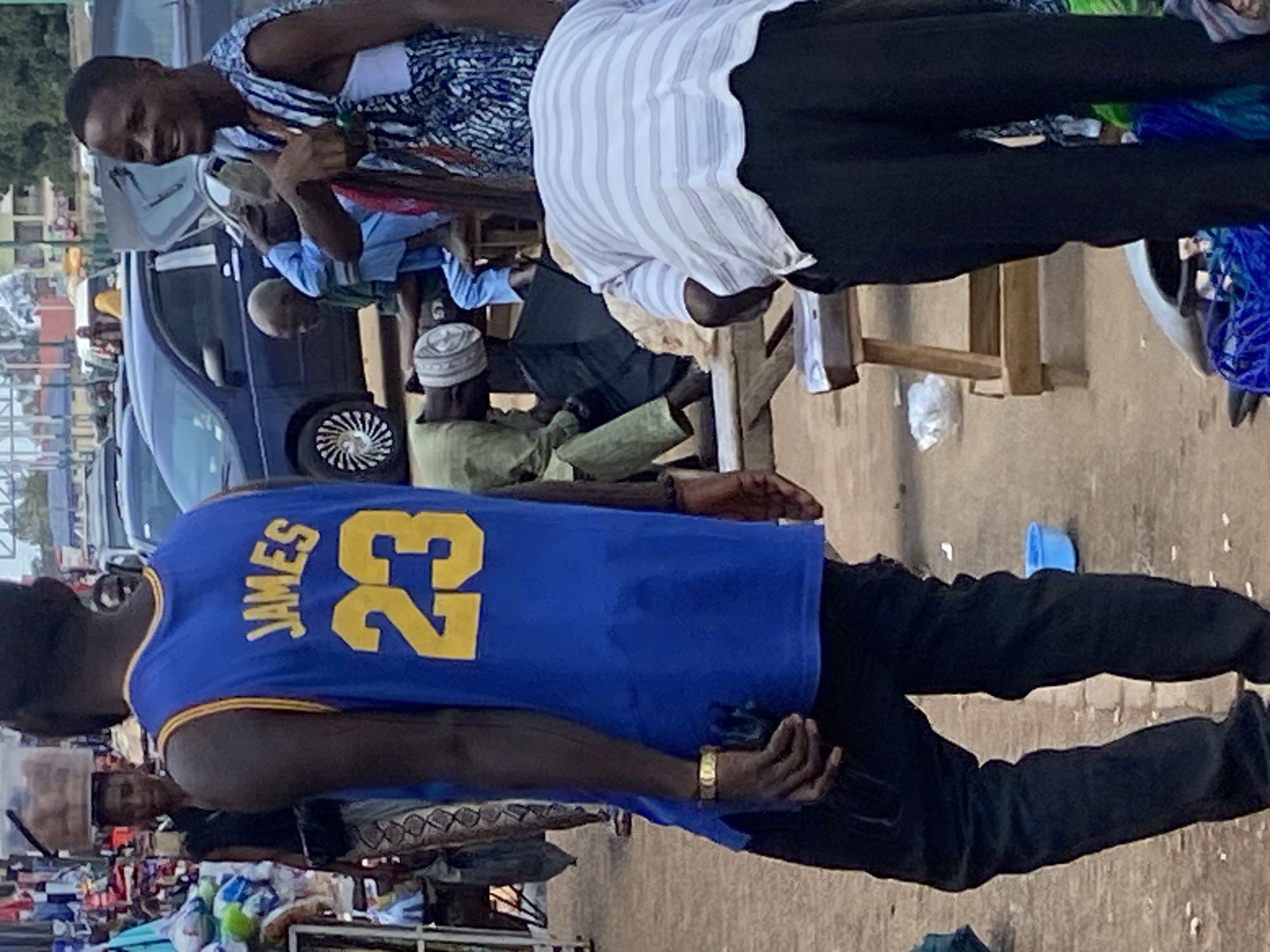 The image depicts a street scene in Bukuru, Nigeria, with several individuals present. In the foreground, a person wearing a blue basketball jersey with "JAMES 23" in yellow lettering faces away from the camera, their back to the viewer. They are wearing black pants and a gold bracelet on their left wrist. To the right, another individual, possibly male, smiles and gestures with their right hand, wearing a patterned sleeveless garment over a white undershirt. Behind these individuals, a group of people are visible, including one person wearing a white skullcap and a green garment, and another in light blue attire. The environment appears to be an outdoor market or roadside setting, with vehicles and stalls in the background, suggesting an urban or semi-urban location. The ground is a dirt or paved surface, with scattered debris and a blue plastic bowl visible on the right. The lighting suggests it is daytime.