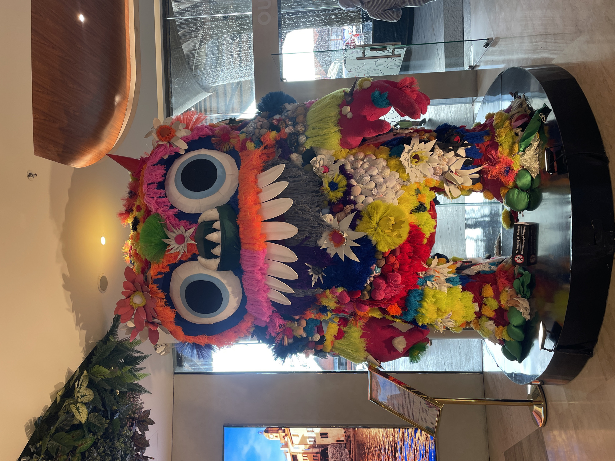 Here's a description of the image:
The image depicts a colorful, artistic monster sculpture displayed indoors. The monster has large, round eyes with blue irises, a mouth with prominent white teeth, and a single red horn. The sculpture is adorned with various flowers, pompoms, and fuzzy materials in a wide range of colors. It stands on a glossy, black platform.
A television screen showing a landscape scene is visible in the foreground. The sculpture appears to be in a modern interior space with large windows and polished floors. There's also a sign on a stand near the sculpture. Additionally, there are decorative green plants to the left of the sculpture.
Based on research, this is likely a "Ogoh-ogoh" sculpture, a demon-like figure built for the Ngrupuk parade on the eve of Nyepi Day in Denpasar, Bali, Indonesia.