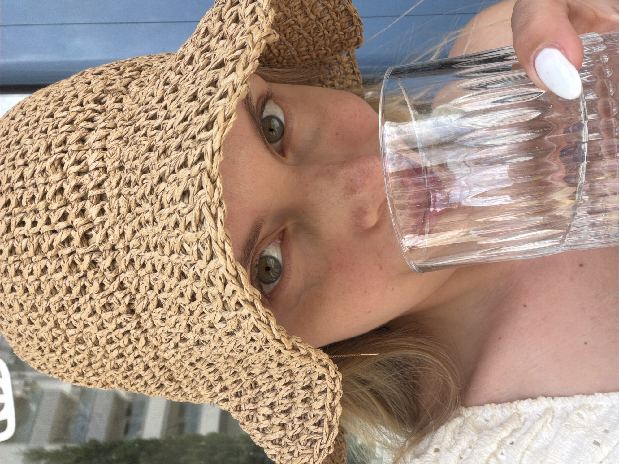 A woman is drinking from a glass in an outdoor setting, possibly in the city of Budva, Montenegro, based on the presence of architectural elements and the outdoor environment. She is wearing a woven straw sun hat that obscures part of her face. Her eyes are visible and appear to be light in color. Her complexion is fair, with some freckles visible on her nose and cheek. She is holding a clear, textured glass to her lips, presumably drinking its contents. She has a white manicure on the hand holding the glass. The visible portion of her clothing appears to be a white, lace-trimmed garment. The background shows blurred architectural details and foliage, suggesting an urban outdoor location.