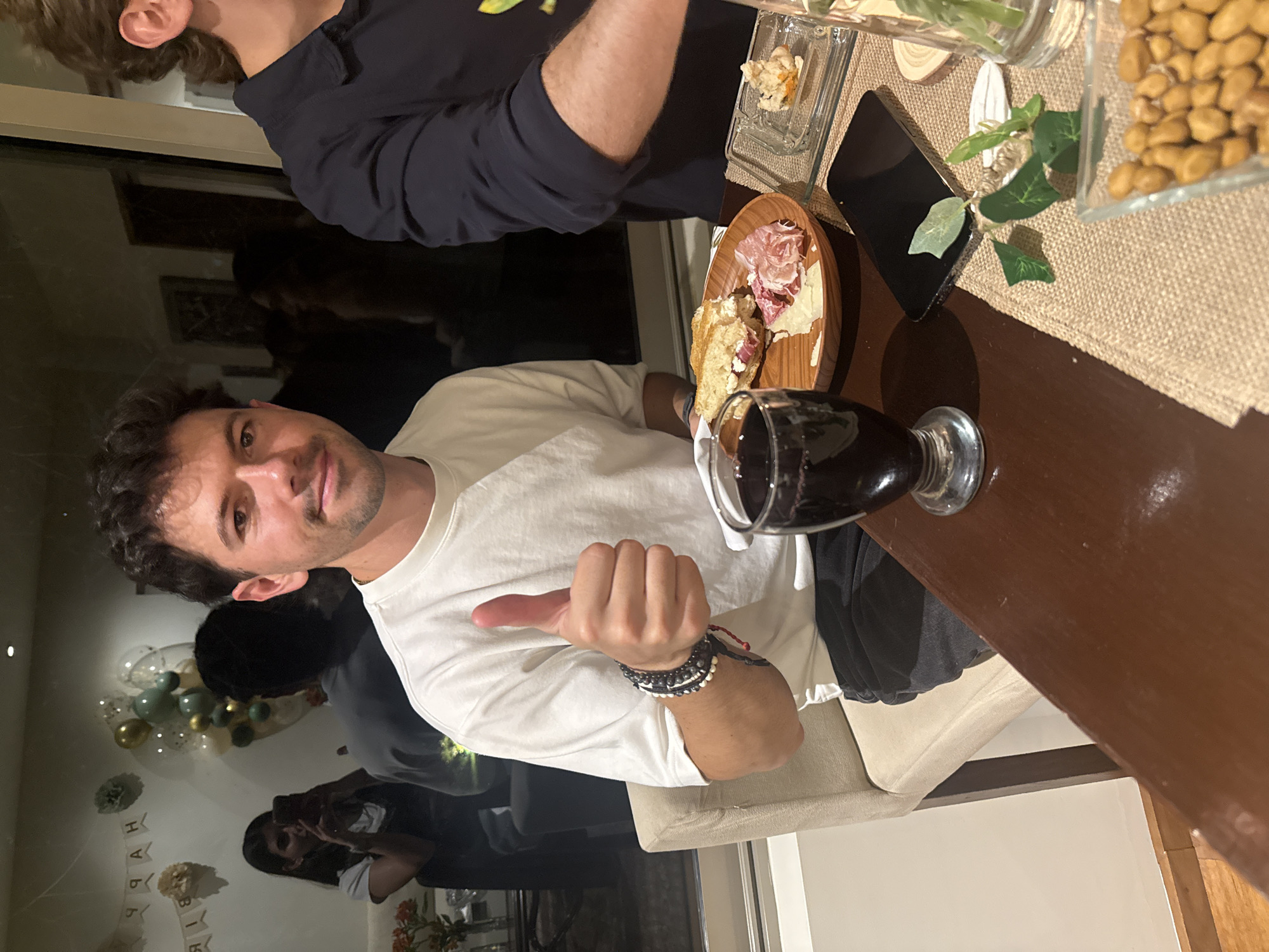 A man in a white long-sleeved shirt is seated at a table with a glass of red wine and a charcuterie board with meats and cheeses. The table is also set with a small appetizer, a smartphone, greenery, and a container of nuts. In the background, there are balloons and a "Happy Birthday" banner. Other people are visible in the background as well. Based on the characteristics of the architecture and the decorations, the location is likely a residence or event space in Mexico City, Mexico.