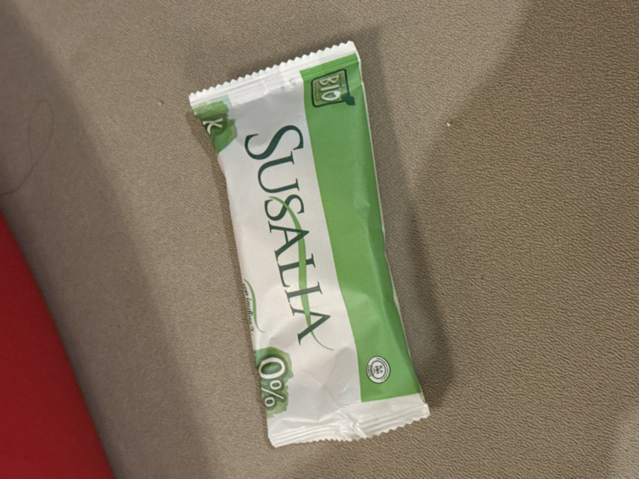 A vertically oriented snack bar, predominantly white and green, rests on a textured tan surface, partially overlapping a red object. The bar features the brand name "SUSALLA" in large, vertically stacked lettering. A "BIO" label is visible at the top, and a "0%" symbol is at the bottom. The wrapper has a crinkled texture and a serrated top edge.