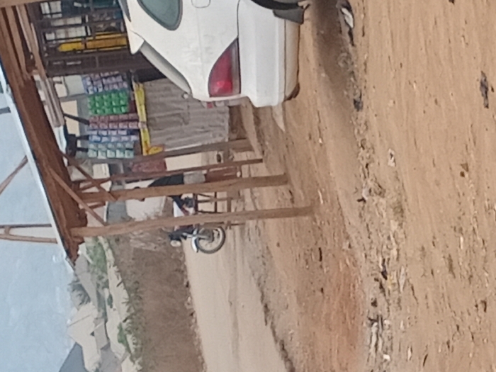 A roadside shop is visible. The shop is a simple wooden structure with shelves stocked with items. A white car is parked partially in front of the shop. A motorcycle is visible near the shop. The area around the shop is a dirt road. The image is taken at a slightly skewed angle.