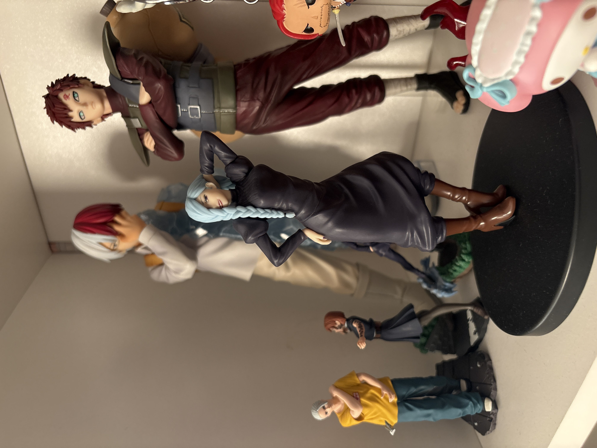 A shelf in a domestic setting displays multiple anime figurines. The figurines include Gaara from Naruto, Todo from My Hero Academia, and an unknown woman in a black dress. Further down the shelf, other figurines are visible but less clear. The backdrop is plain and light-colored, suggesting a modern interior. The location, Plovdiv, Bulgaria, is not directly discernible from the image.