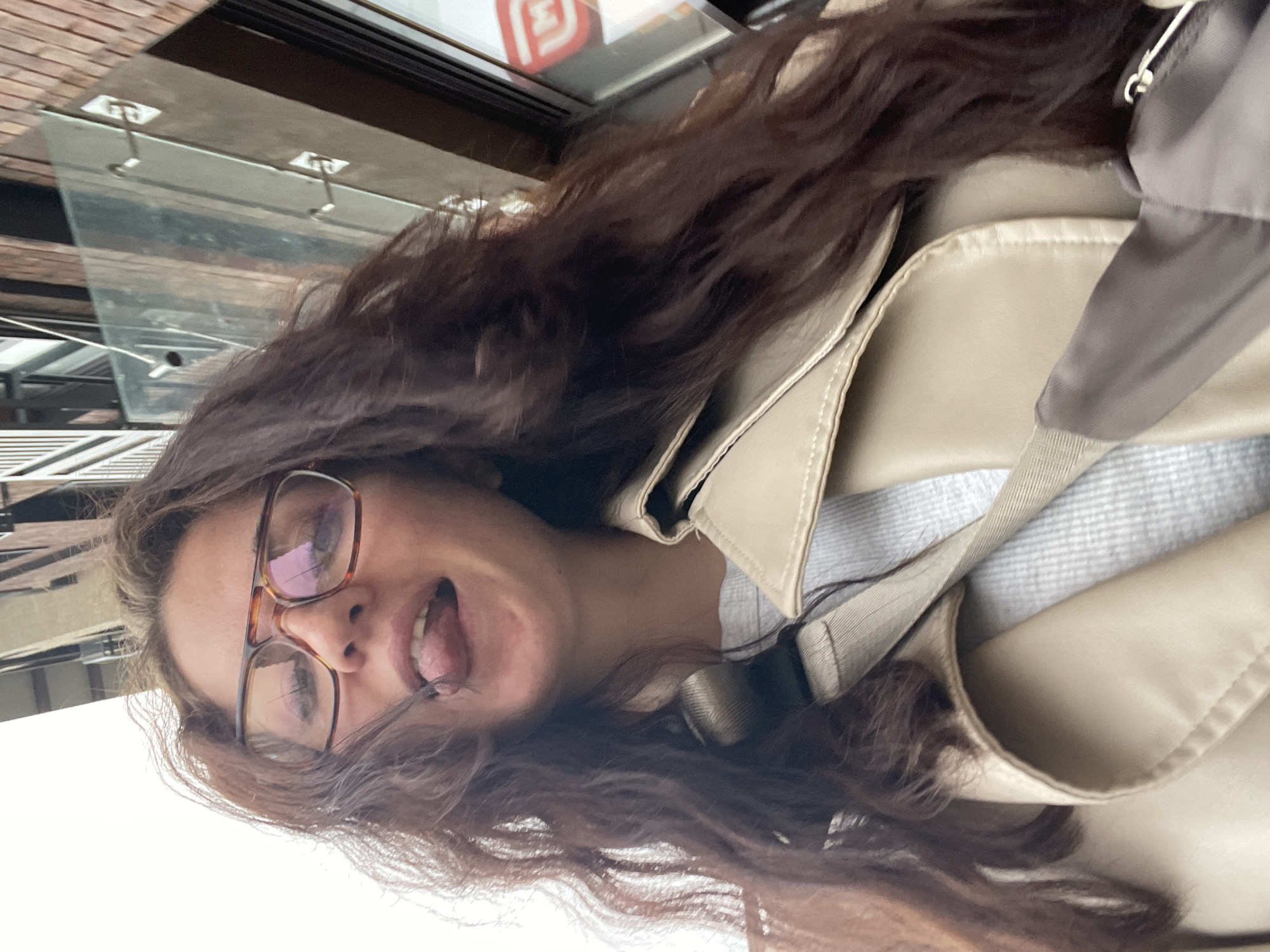 A woman with long, brown, wavy hair and glasses is seen in an eye-level, tilted, outdoor shot. She is wearing a tan trench coat and a light grey patterned shirt. Her tongue is sticking out. In the blurred background, there's a building with a glass awning, suggesting an urban environment. The city of Razvilka, Russia is not discernible from the image.