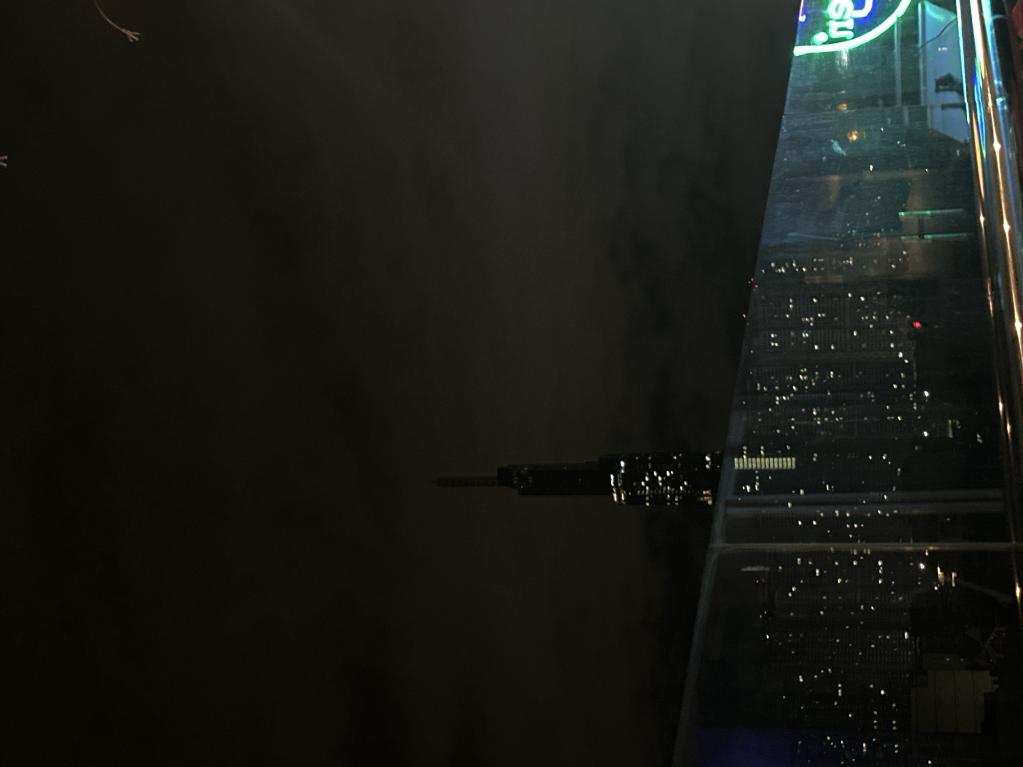 The image captures a nighttime cityscape from a high vantage point, likely a rooftop or a tall building. In the background, a prominent, slender skyscraper dominates the skyline, illuminated by numerous lights, suggesting it is a modern architectural marvel. The sky is dark and overcast, with some faint glows indicating urban light pollution. In the foreground, a glass barrier reflects the city lights, creating a blurred, shimmering effect. The city itself appears densely populated with many buildings adorned with lights, creating a vibrant, twinkling panorama. A neon sign with blue and green lights, partially visible on the right, adds a touch of contemporary advertising to the urban scene. The overall impression is that of a bustling metropolis at night, with the dark sky contrasting against the artificial lights of the city. The presence of the specific neon sign suggests a commercial or entertainment district within Thủ Đức, Vietnam.