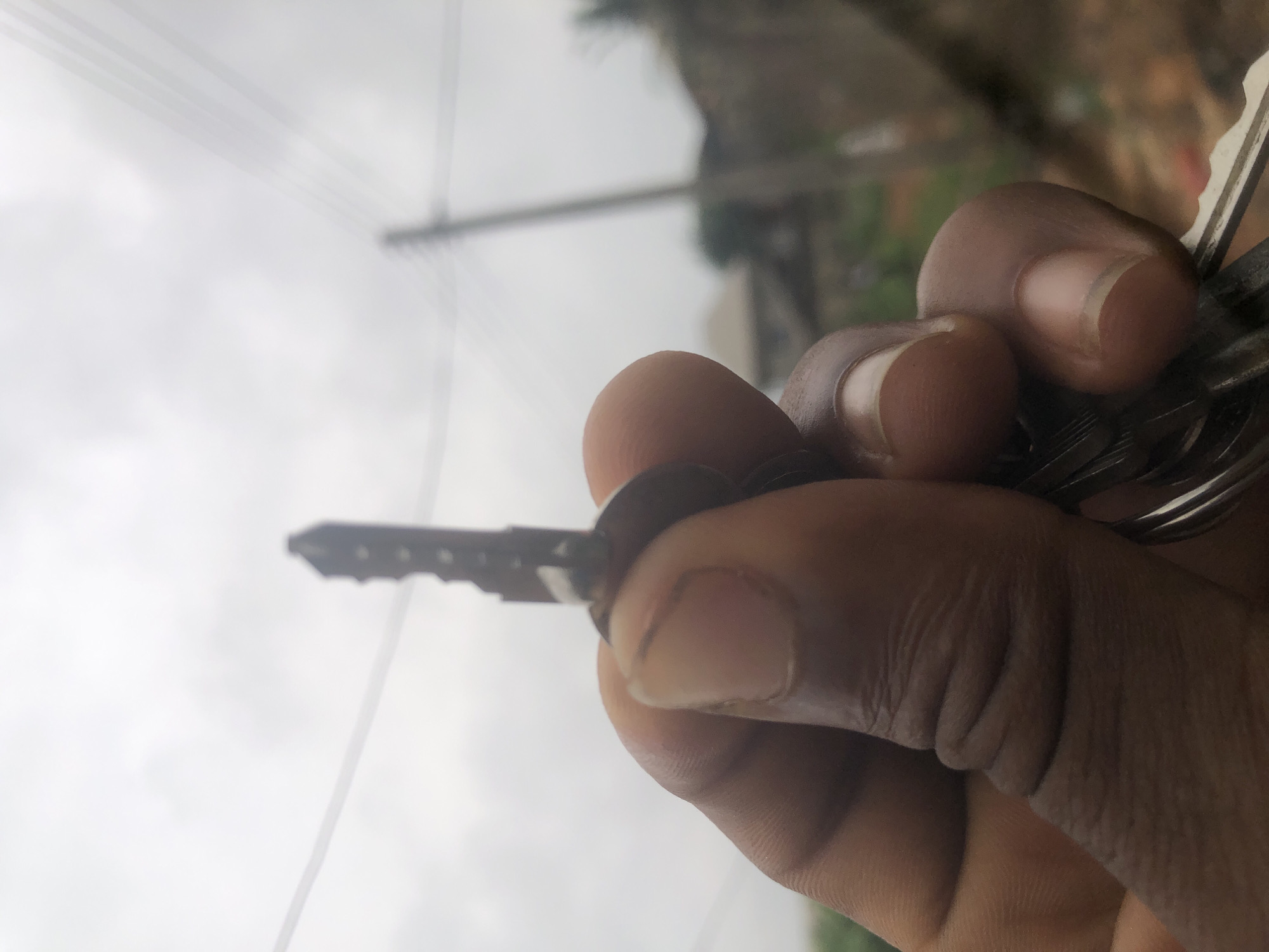 The image presents a close-up, vertically oriented shot of a human hand holding a set of keys against a heavily blurred background. The hand has a dark skin tone, with fingers and thumb clearly visible as they grip the keys. One specific key, dark in color with a distinctive jagged-edged blade, is prominently held between the thumb and forefinger, extending towards the left of the frame. Other keys on the keyring are partially visible, clustered behind the main key.
The background is significantly out of focus, creating a shallow depth of field that emphasizes the hand and keys. It consists primarily of a bright, overcast sky, indicating daytime conditions. Faint, dark lines resembling power lines or cables are discernible stretching across the upper left portion of the blurred sky. A dark, vertical shape, possibly a utility pole or similar outdoor structure, is vaguely visible in the upper right background. There are also very indistinct hints of green, suggesting distant foliage or trees, further contributing to an outdoor setting.
The activity depicted is the simple act of holding keys. No specific event or emotion is discernible beyond this. There is no visible text within the image. The scene appears to be an ordinary outdoor setting, likely urban or suburban, under cloudy or overcast weather conditions.