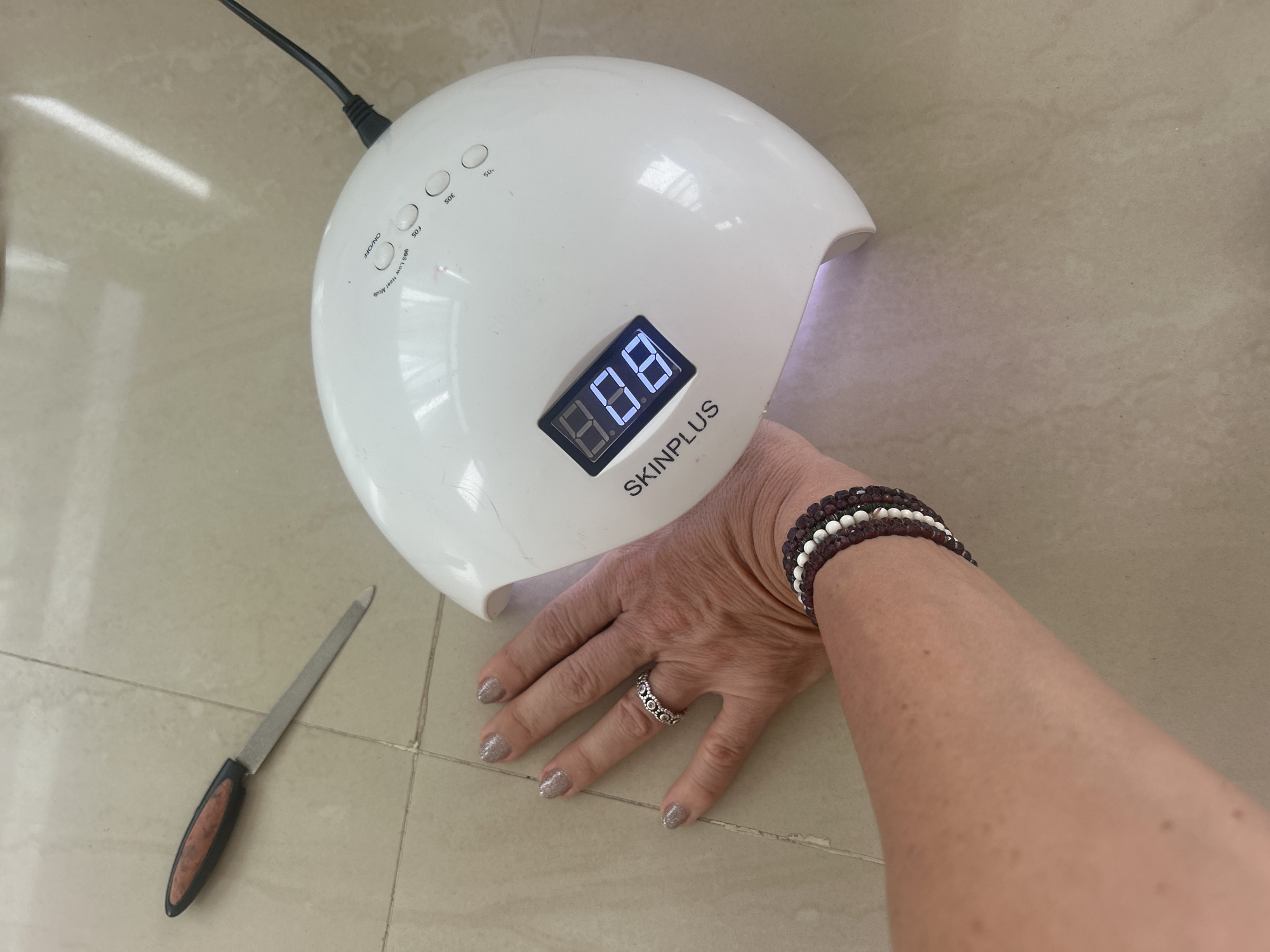 The image shows a woman's hand being held under a white, dome-shaped UV nail lamp on a tiled surface. The lamp displays "8.8.8" on its digital timer and is labeled "SKINPLUS." To the left of the lamp, a metal nail file with a black and brown handle is visible. The woman is wearing a silver ring and beaded bracelets. The location is Puttaparthi, India.