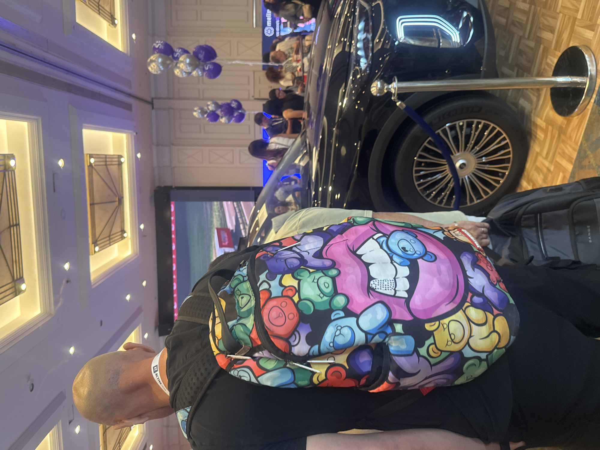 The image shows a man with a bald head wearing a black t-shirt and black pants, carrying a backpack with a colorful design featuring teddy bears and lips. He is standing in an indoor venue, possibly a convention center or exhibition hall, with a large screen displaying an image in the background. A black car is parked behind him, and a crowd of people are visible in the distance. Balloons are also present, some of which appear to be branded. The floor is covered in a wooden parquet pattern. There is no indication that the location is in Tel-Aviv, Israel.