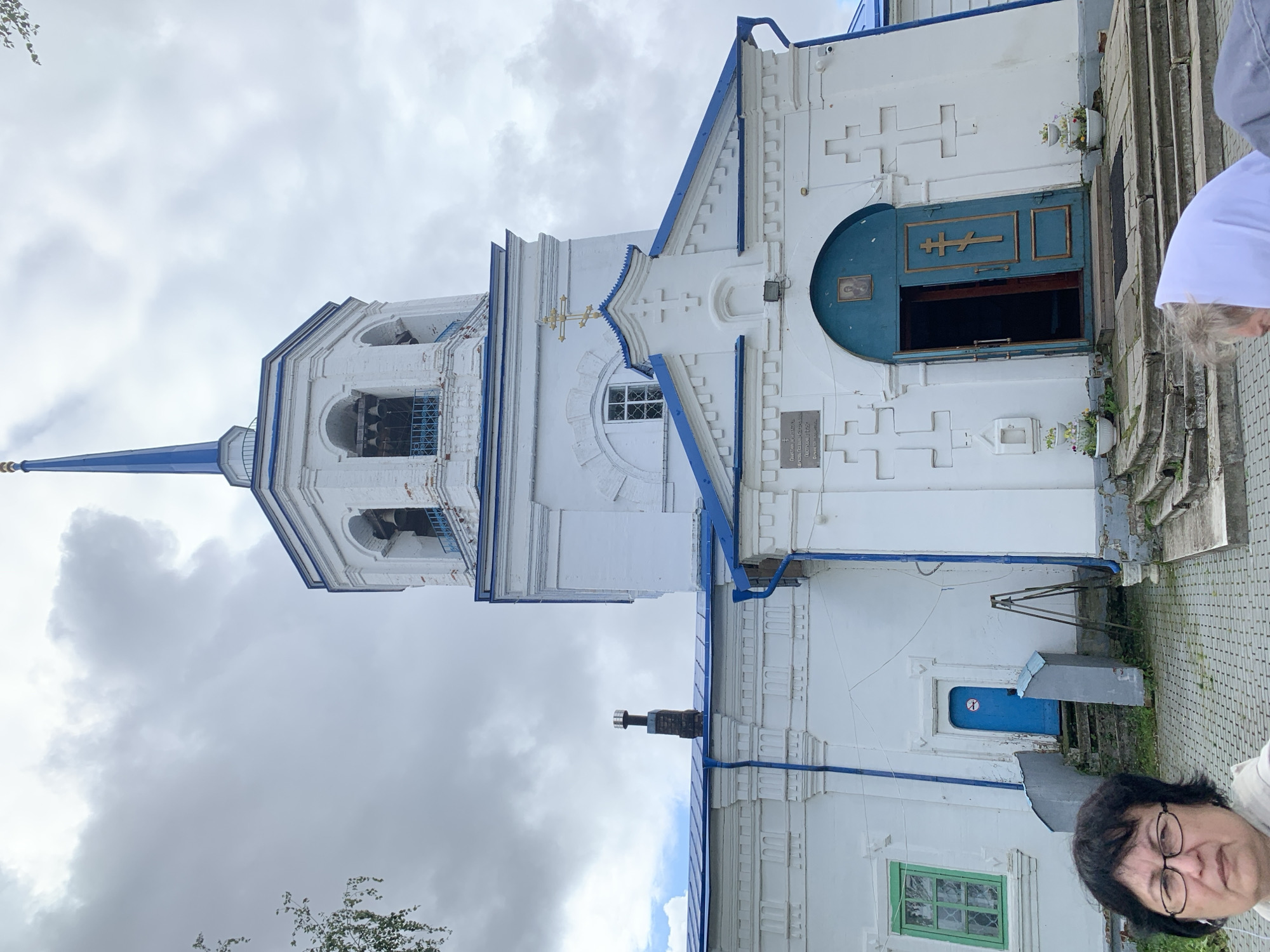 The image shows a white church with a blue roof and a tall bell tower topped with a blue spire. The church has several windows, some with green frames, and a large blue double door with a golden cross. There are two women in the foreground, one looking at the camera and another bending over. The ground is paved with cobblestones. The sky is cloudy. The church is located in the city of Oryol, Russia. The specific location is the Church of the Resurrection of Christ, located at ul. Pokrovskaya, 100.