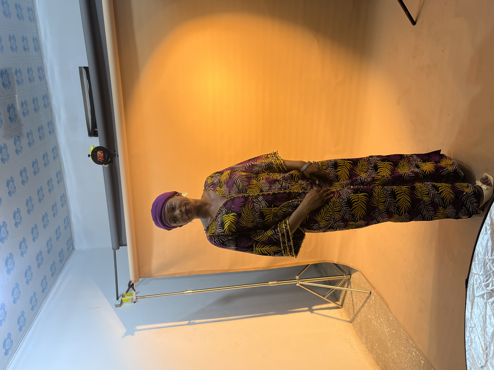A woman stands indoors against a yellow-beige backdrop. She wears a purple head covering and a dark patterned dress with yellow palm leaf designs. Her hands are clasped in front of her. Behind her is a photography lighting setup, including a silver reflector. The room has a patterned blue-and-white wall, indicating an interior space. The setting appears to be a studio in a home. Gura Top, Nigeria, is not definitively recognizable from this image.