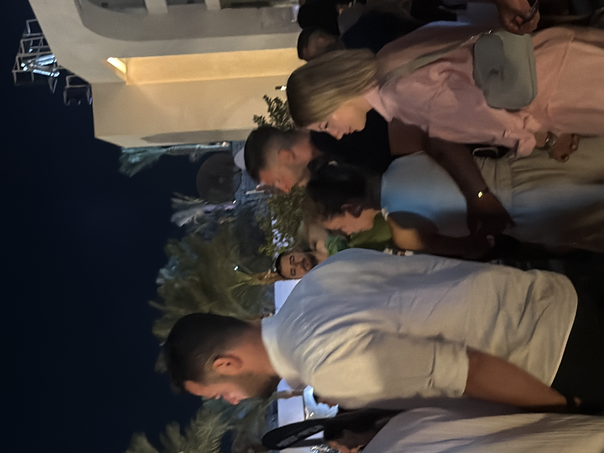 The image depicts a nighttime outdoor gathering in Sharm El Sheikh, Egypt. In the foreground, a man in a white button-down shirt with rolled-up sleeves stands in profile facing left, his hand in his pocket. Behind him and to the right, a woman in a light pink outfit with a light blue crossbody bag is also facing left. Several other individuals are visible in the background, some in profile, engaged in conversation or observing their surroundings. Palm trees and architectural elements suggestive of a resort or venue are present in the dimly lit environment. The overall scene suggests a social event or a casual get-together.