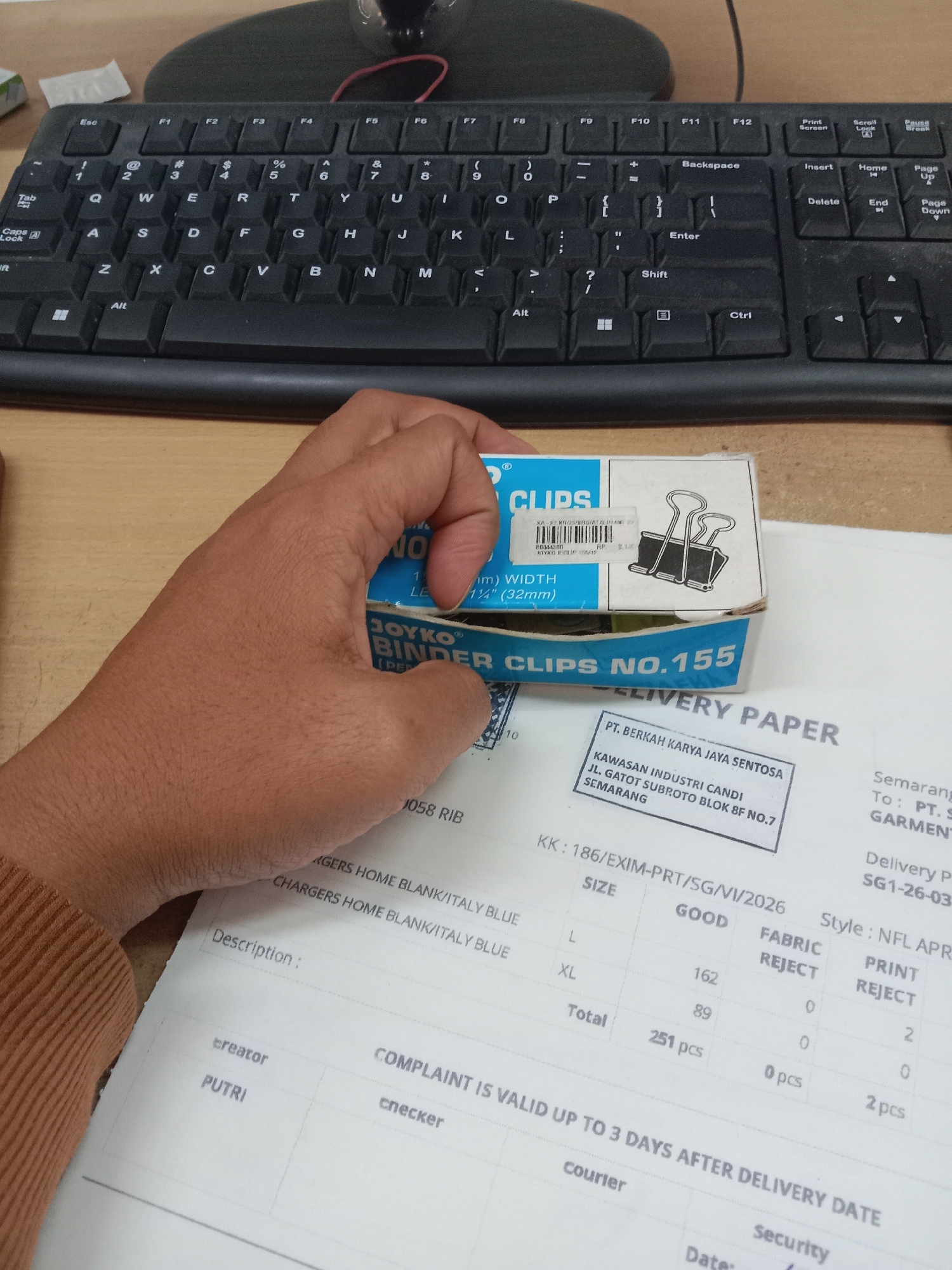 An overhead medium shot captures a person's right hand holding a box of "JOYKO" binder clips, with the clips partially visible inside. The hand is positioned above a "DELIVERY PAPER" that is spread out on a desk, suggesting a transaction or inventory process. The paper contains printed text and a stamp with the address "JL. GATOT SUBROTO BLOK 8F NO.7 SEMARANG", indicating the location is in Semarang, Indonesia. The desk also features a black computer keyboard and a portion of a monitor base, implying an office or workspace environment. The lighting is somewhat dim, casting soft shadows.