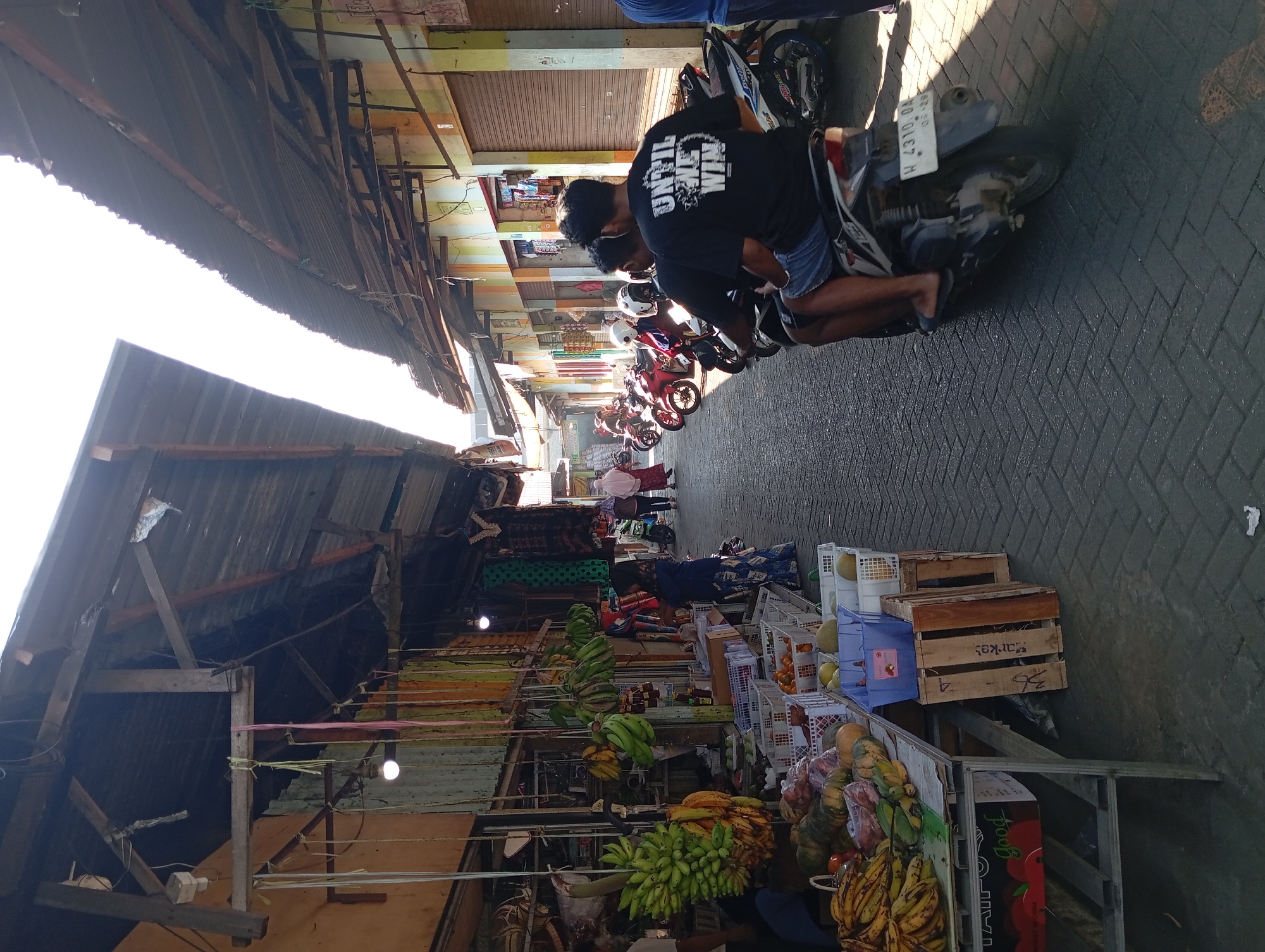 This is a photograph of a street market in Kepulungan, Indonesia, under a bright sky. The street is paved with interlocking bricks. On the left side of the street, there are market stalls displaying various fruits, including bananas and pumpkins. Colorful fabrics hang from the awnings of the stalls. On the right side of the street, there are shops with closed metal shutters. In the middle of the street, several motorcycles are parked. Two men are riding a motorcycle, with one man in the foreground wearing a black t-shirt with white lettering that reads "UNTIL WE WIN." Other people, including women in headscarves, are walking in the background. The overall scene depicts a lively and active market environment.