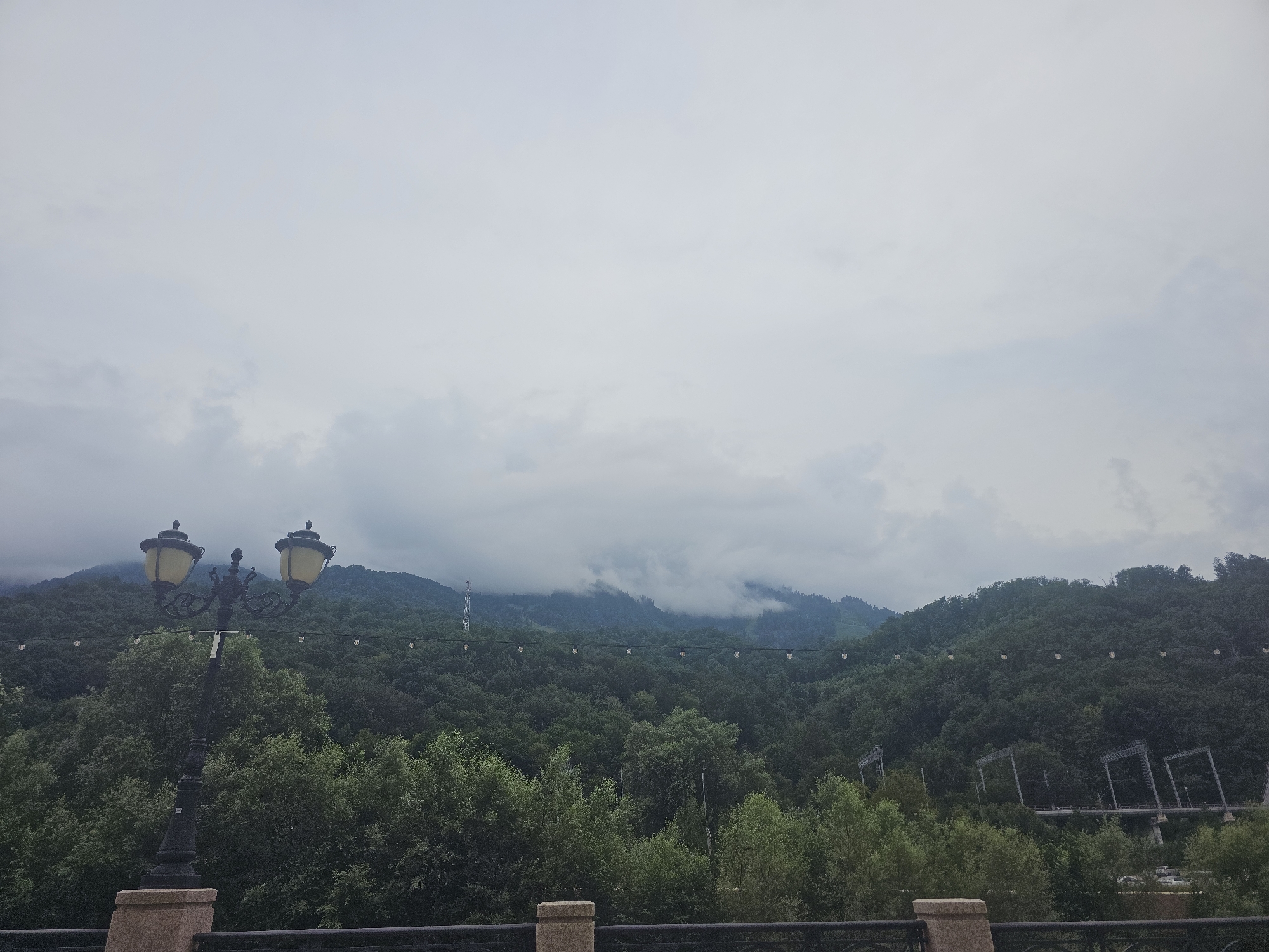 This is a landscape photograph taken in the city of Estosadok, Russia. The scene depicts a view of a lush, tree-covered mountain range under a cloudy sky. In the foreground, there is a decorative lamppost with two lights, and a low stone railing with a metal fence. String lights are visible stretching across the frame, suspended in front of the trees. In the background, the mountains are shrouded in mist, with some structures, possibly part of a ski resort or infrastructure, visible on the slopes. The overall impression is one of a natural landscape with some man-made elements integrated into it, likely indicating a tourist or recreational area.