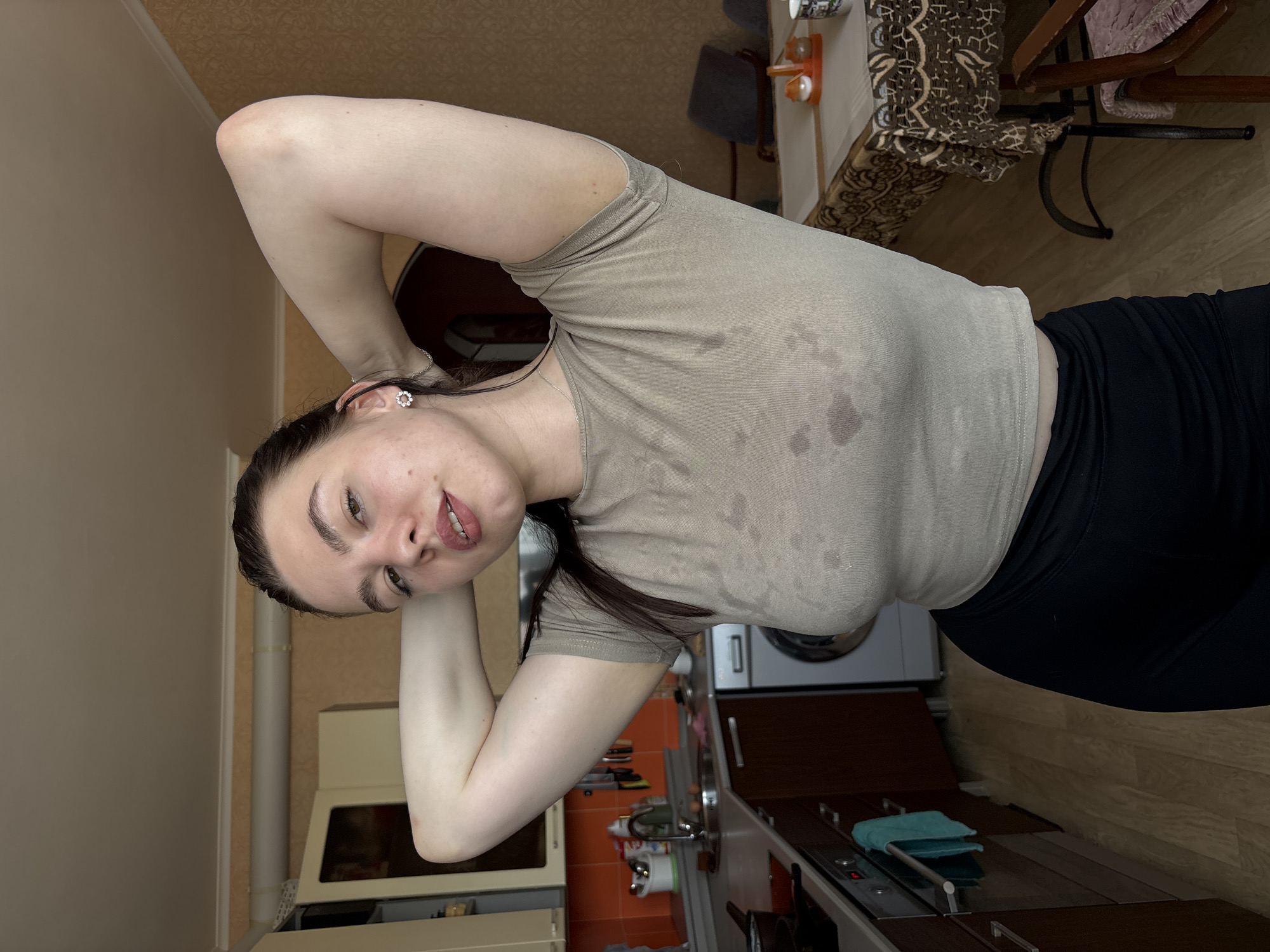 The image shows a woman in a kitchen, likely in an apartment. She is wearing a beige t-shirt and black leggings. Her hair is dark, and she has her hands behind her head. The kitchen contains typical appliances and cabinets. The location is suggested to be Perm, Russia, based on external information, but there are no details in the image to confirm this.