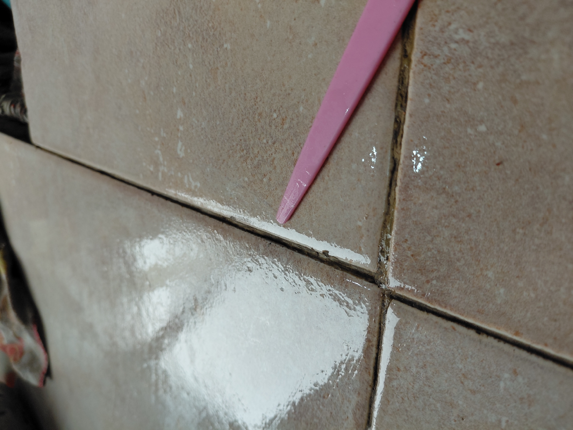 The image displays a close-up, angled view of beige-brown ceramic tiles with dark grout lines, possibly in an interior setting such as a bathroom or kitchen in Santa Ana, Venezuela. No people are visible.
The scene is dominated by two rows of rectangular tiles, one above the other, separated by a horizontal dark grout line. A vertical dark grout line divides the lower tiles. The tiles have a slightly textured surface and vary in shade, exhibiting lighter speckles or residue in several areas.
A slender, pink plastic tool, possibly a scraper or spatula, is angled across the upper horizontal grout line, its tip resting against the tile surface. This suggests an ongoing activity involving cleaning, scraping, or applying a substance to the grout or tiles.
A notable detail is the significant amount of wetness or a glossy clear substance covering portions of the tiles and within the grout lines, creating reflections, particularly prominent on the lower left tile. This wetness could be water, a cleaning solution, or a sealant. The wet areas reflect light, giving them a shiny appearance. Some of the grout appears to have white or light-colored residue along its edges, contrasting with its dark color. No visible text is present in the image.