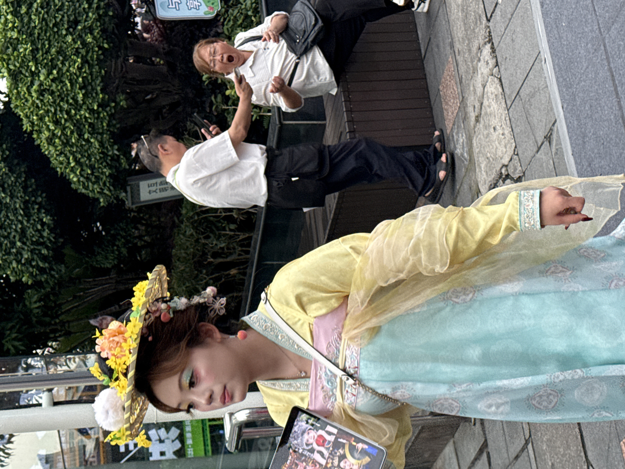 In Yuzhong District, China, a young woman adorned in a traditional yellow and blue garment, featuring ornate embroidery and a floral headdress, stands on a paved walkway. She holds a tablet in her left hand, displaying images. To her right, a man in a white shirt and black trousers is seen holding a mobile phone, with his hand extended towards a seated woman. The woman, wearing glasses and a striped shirt, has her mouth wide open in a yawn or shout, and her hands are positioned as if in surprise or exertion. She is carrying a black handbag. Behind them, lush green trees and foliage form a backdrop, with a wooden bench visible where the woman is seated. The scene suggests an outdoor urban environment, possibly a street or a park area.