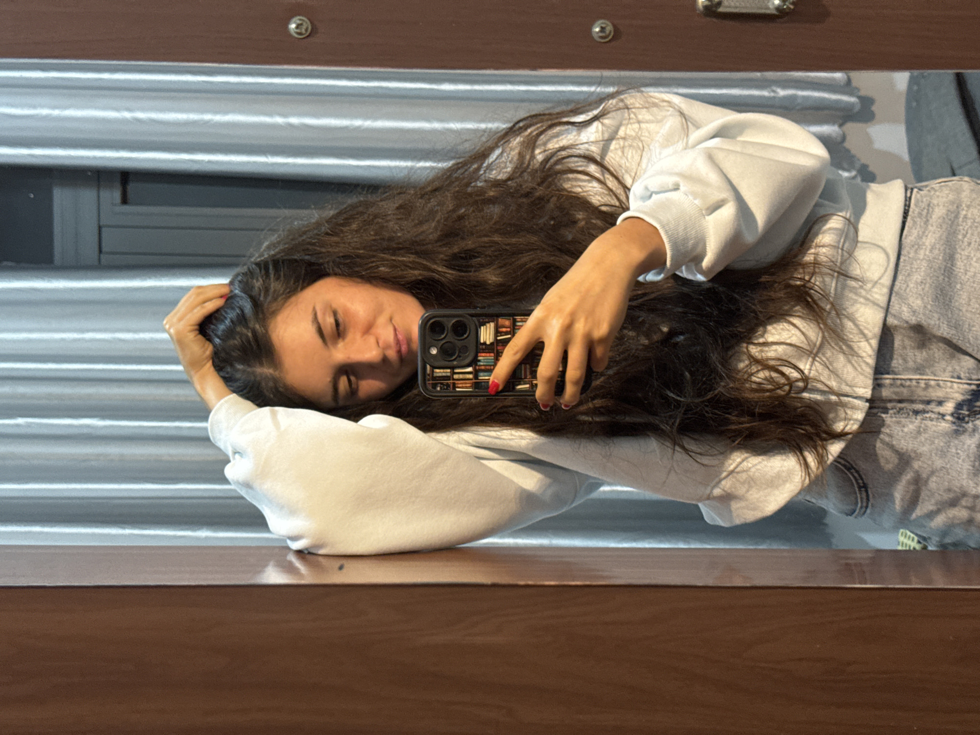 A young woman with long, dark, wavy hair, wearing a light blue hooded sweatshirt and denim shorts, is taking a mirror selfie. Her left arm is raised, with her hand resting on top of her head, and her right arm is holding a smartphone with a colorful, patterned case. She is leaning against a brown wooden doorframe. The background features a sheer, light-colored curtain. The scene appears to be indoors, likely in a residential setting, and there is no indication that the location is in Mũi Né, Vietnam.