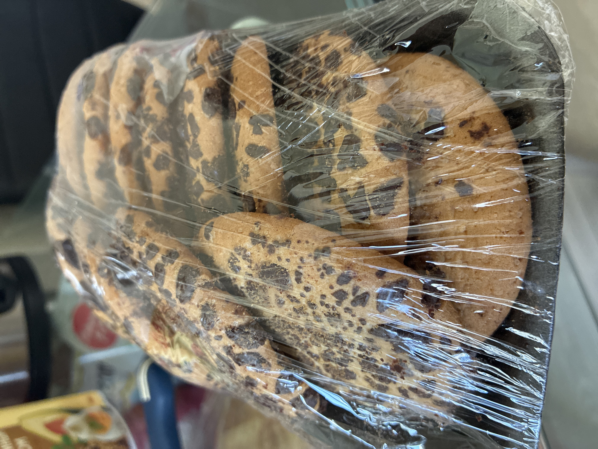 The image shows a close-up view of a package of chocolate chip cookies. The cookies are round and appear to be freshly baked, with visible chocolate chips throughout. They are arranged in a tray and covered in clear plastic wrap, which is slightly crinkled. The lighting suggests an indoor setting, possibly a store or a kitchen. The background is out of focus, but hints of shelves and other products can be discerned, indicating a commercial environment. No people are visible in the image. The focus is entirely on the cookies, highlighting their texture and chocolate distribution. The scene is static, with no particular activity taking place other than the presentation of the product. There is no visible text in the image. The location context provided is Gornye Kljuchi, Russia, but there are no visual cues within the image to confirm this specific location.