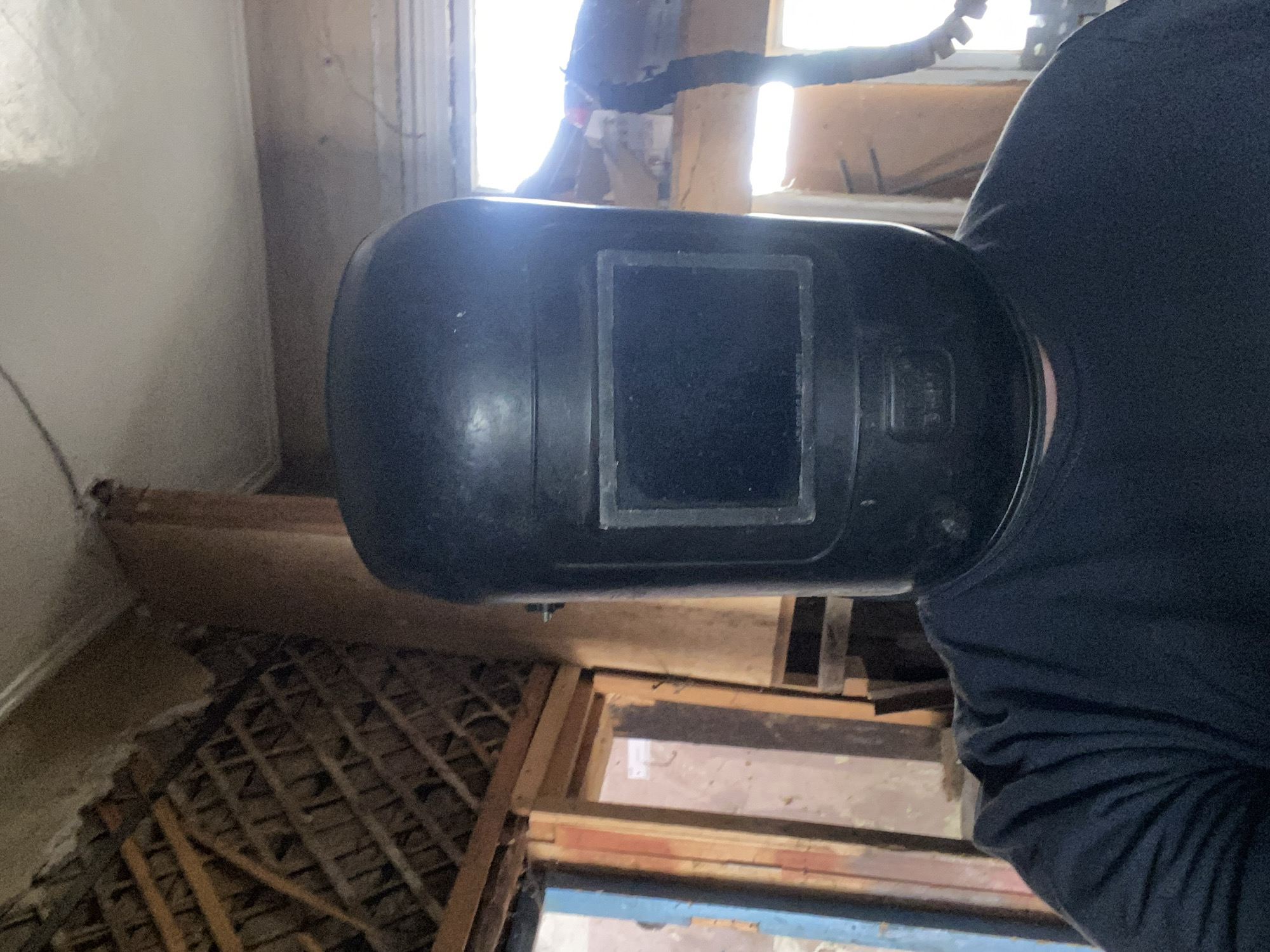 In Oktyabrsk, Russia, a person wearing a black welding helmet and a black t-shirt stands in what appears to be an unfinished or dilapidated interior space. The background shows exposed wooden beams and rafters on the ceiling, suggesting construction or disrepair. To the left, there are openings that resemble doorways or windows, with visible wooden framing and panes of glass. Light streams in from the right side of the image, indicating an external light source. The overall environment is dimly lit, with a focus on the subject in the foreground.