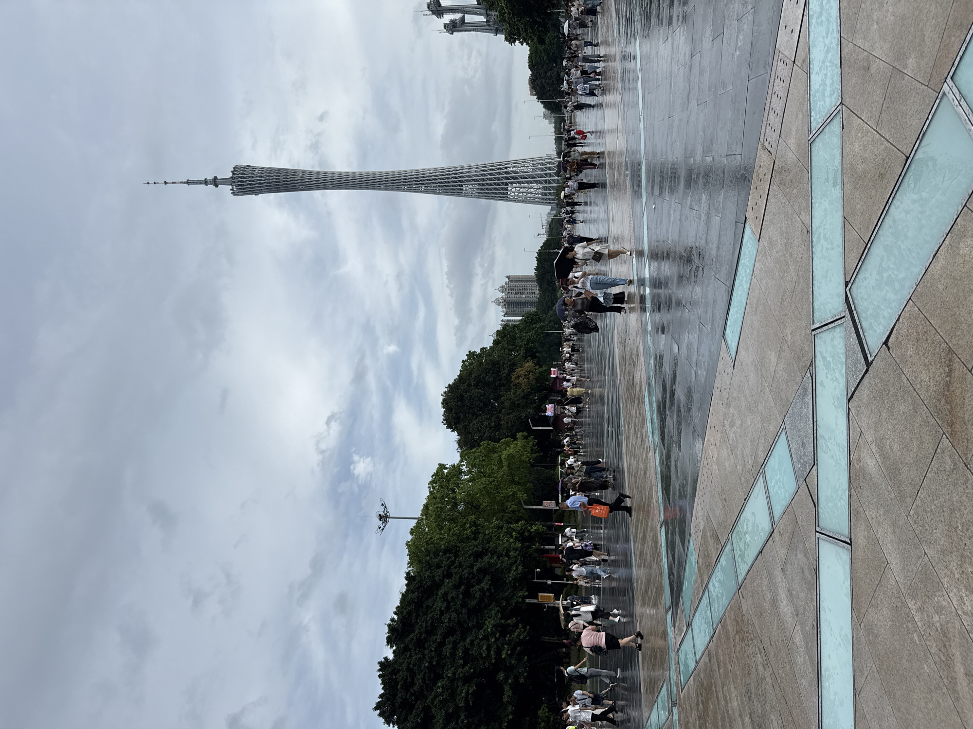 The image shows a wide, wet, paved plaza in front of the Canton Tower, located in the Tianhe District of Guangzhou, China. The sky is overcast with gray clouds. Numerous people are walking across the plaza, some carrying umbrellas, suggesting it has recently rained or is still drizzling. The ground is reflective due to the moisture. In the foreground, the plaza features geometric patterns of stone paving with embedded strips of frosted blue glass or light. The Canton Tower, a distinctive lattice-like observation tower, dominates the background, towering over the scene. Trees line the sides of the plaza, and several other modern buildings and structures are visible in the distance.