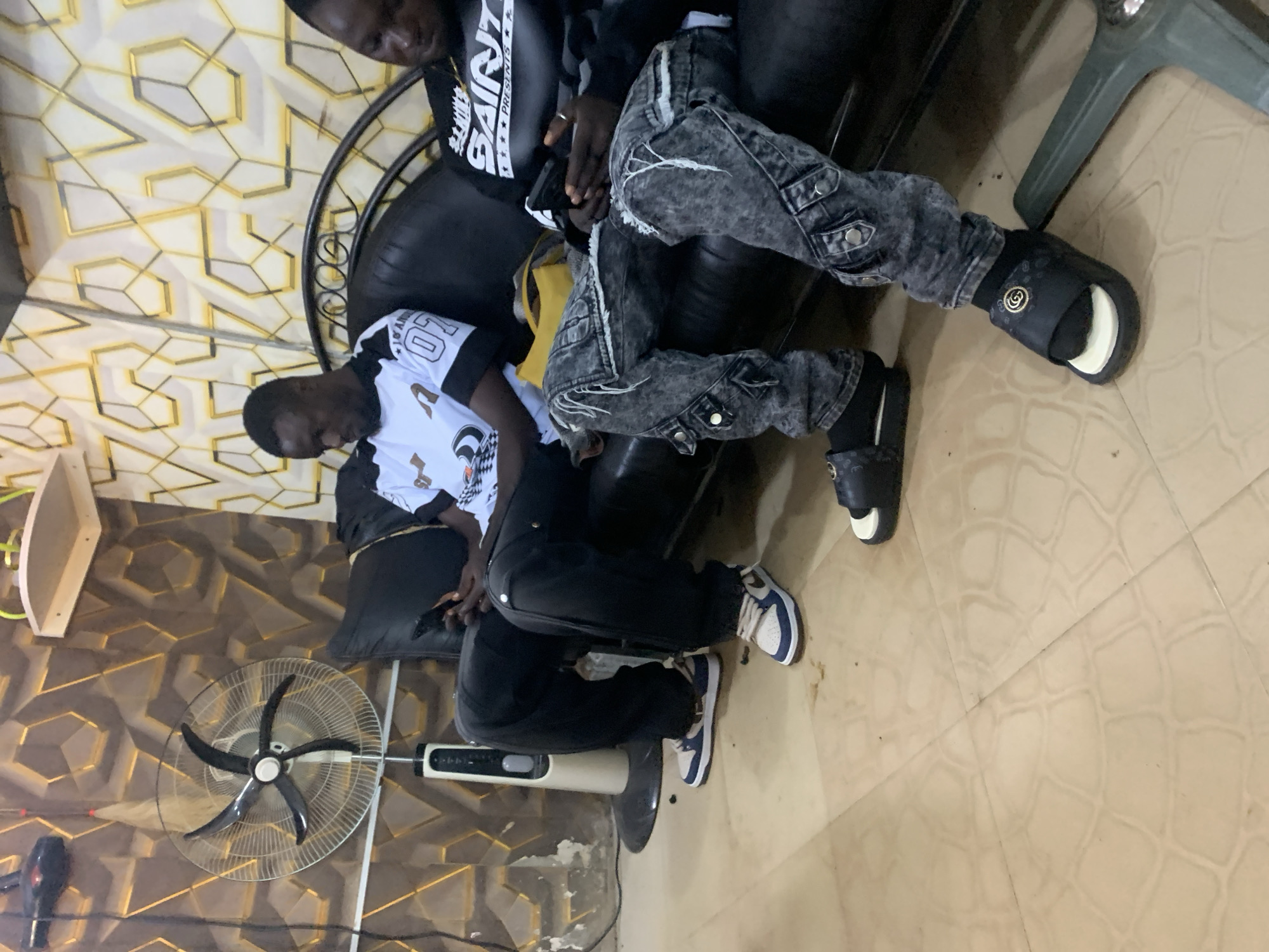 Two young Black men are seated on a black leather sofa in an indoor room. The man on the left is wearing a white t-shirt with black sleeves and graphics, black pants, and blue and white sneakers. He is holding a cell phone in his hands. The man on the right is wearing a black t-shirt with graphics and distressed, dark wash cargo pants, along with black socks and black slide sandals. He is also holding a cell phone. Behind them, the wall features a geometric pattern with gold accents. To the left of the sofa, a white pedestal fan stands. The floor is tiled in a light tan color with a circular pattern. The scene appears to be a casual indoor setting, possibly a lounge or waiting area.