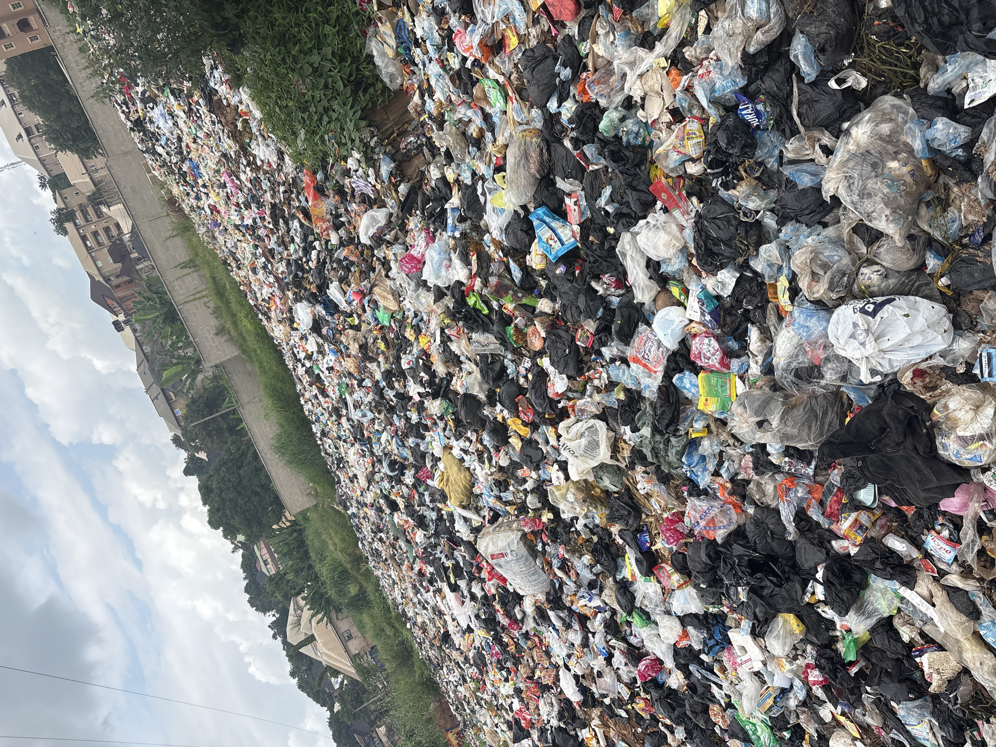 This media file captures a vast expanse of illegally dumped waste, predominantly plastic bags and packaging, piled high along a roadside in Nkpor, Nigeria. The scene is overwhelming, showcasing the sheer volume of refuse that has accumulated. Amongst the debris, a variety of objects are discernible: white and black plastic bags, colorful wrappers, bottles, and discarded containers. The garbage is densely packed, creating an uneven and chaotic landscape.
To the left of the dump, buildings and trees are visible, suggesting an urban or semi-urban setting. The presence of these structures provides a stark contrast to the environmental degradation. The image appears to have been taken during the daytime, with diffused light suggesting it could be overcast or late afternoon. The sky, visible in some crops, is cloudy.
There is no discernible activity or event taking place beyond the existence of the dump itself, implying a static, long-term problem. The overall impression is one of neglect and environmental crisis. Some faint text is visible on packaging, such as "PURITY" on a blue and white packet, and "ipo" on a red and white packet. However, much of the text is obscured or illegible due to the nature of the waste. The scale of the dump and the surrounding environment hint at the significant challenges faced by communities in managing waste in this location.