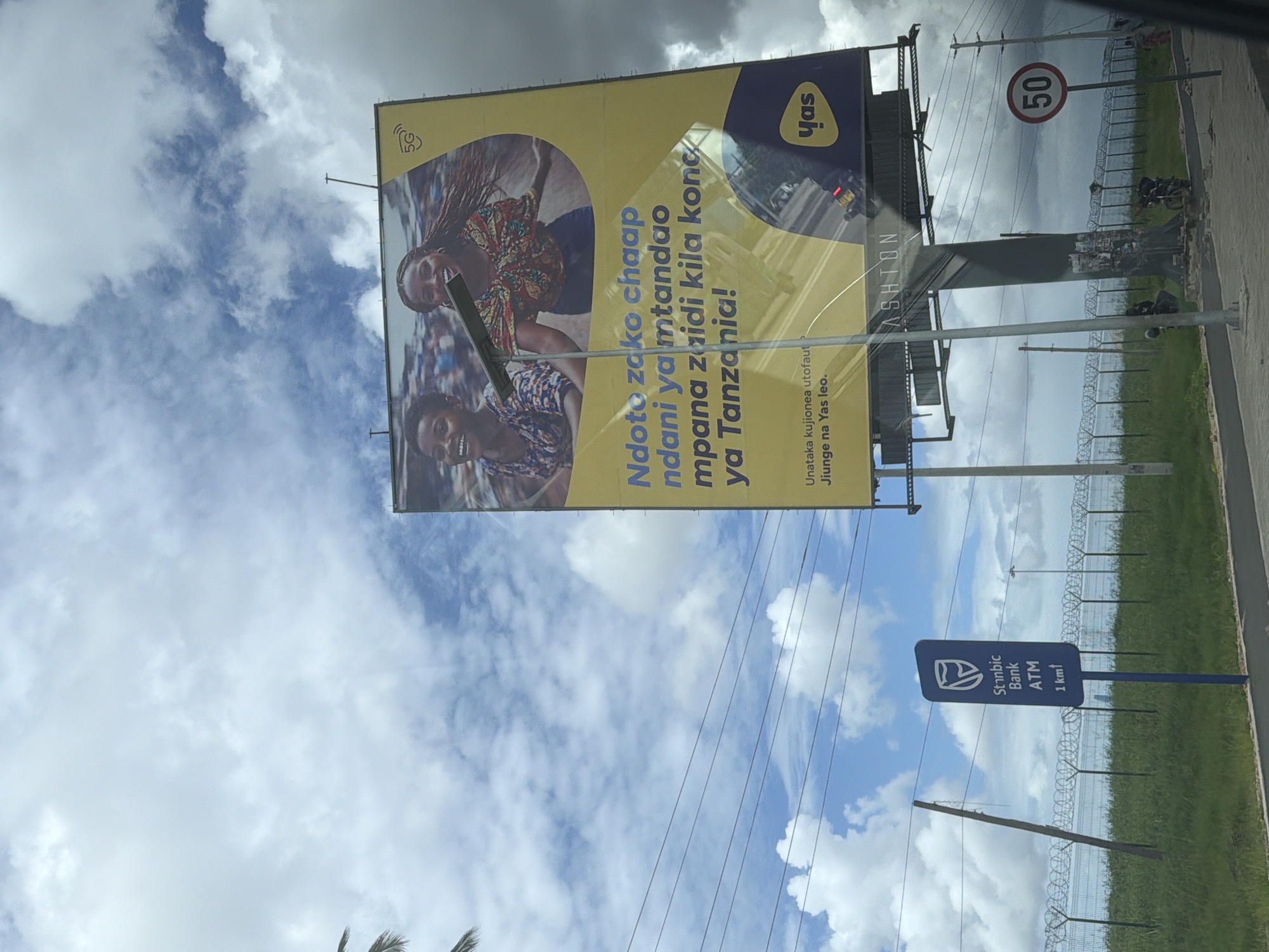 The image shows a large advertising billboard with the YAS logo and a promotional image of smiling people. The advertisement includes the Swahili text "Ndoto zako chaap ndani ya mtandao mpana zaidi kila kona ya Tanzania!" It also says "Unataka kujionea utofauti... Jiunge na Yas leo." The billboard is situated above a sign indicating a Stanbic Bank ATM is 1 km away. In the background, there is a fence with barbed wire, green foliage, a "50" speed limit sign, and partly cloudy sky. The image appears to be taken outdoors, possibly in a developing country, but the specific location in the city of Unknown, Unknown is unknown.