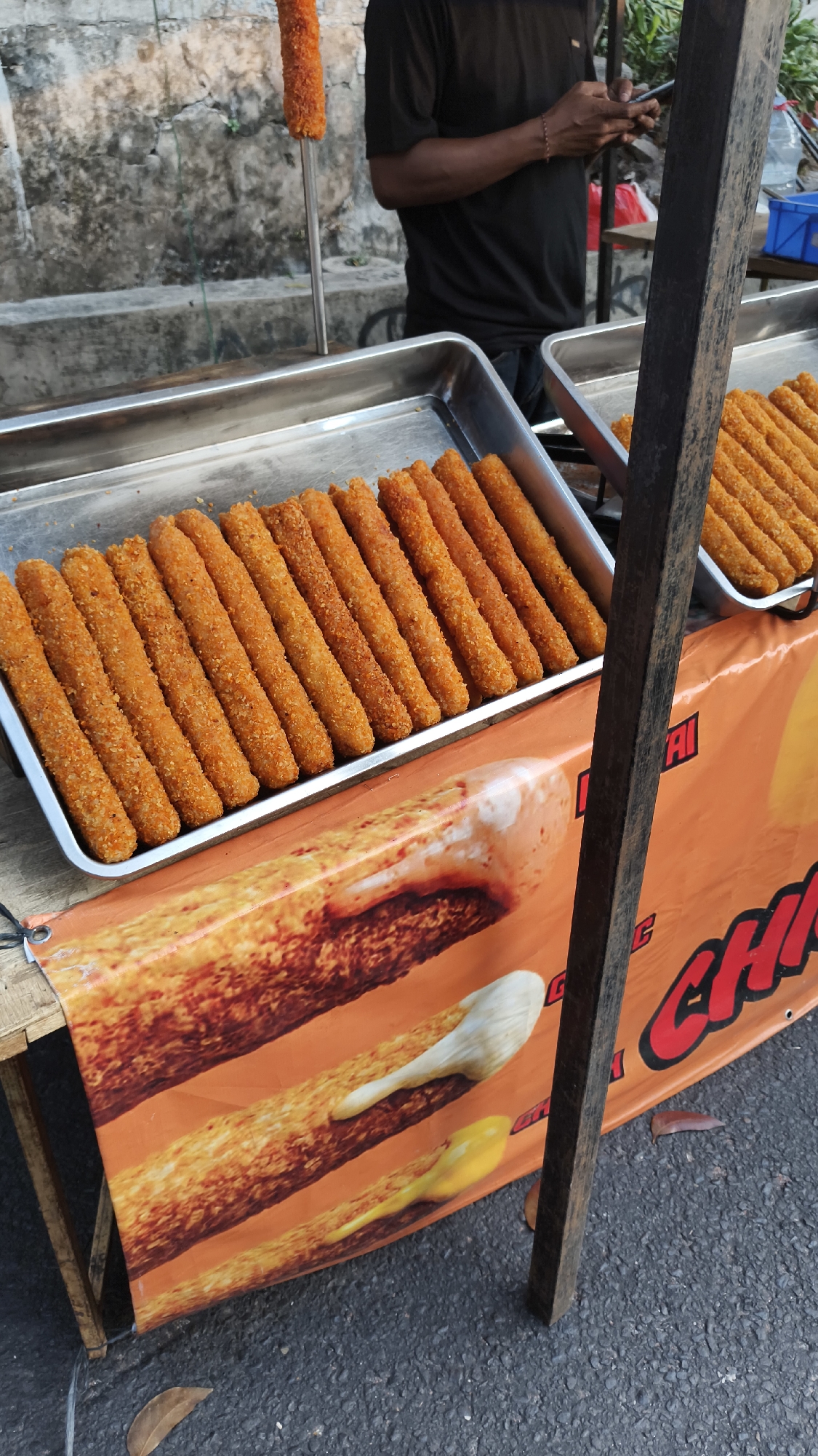A street vendor in South Jakarta, Indonesia, is seen standing behind a cart laden with fried snacks. The cart features a banner with images of large fried snacks, some with cheese and sauce. In the foreground, a tray holds numerous golden-brown, breaded fried sticks, likely some form of snack food. Another tray with similar snacks is visible in the background. The vendor, wearing a black t-shirt, appears to be interacting with a mobile phone. The environment suggests an outdoor setting, possibly a market or a busy street, with a textured wall in the background and asphalt on the ground. The lighting indicates it is daytime.