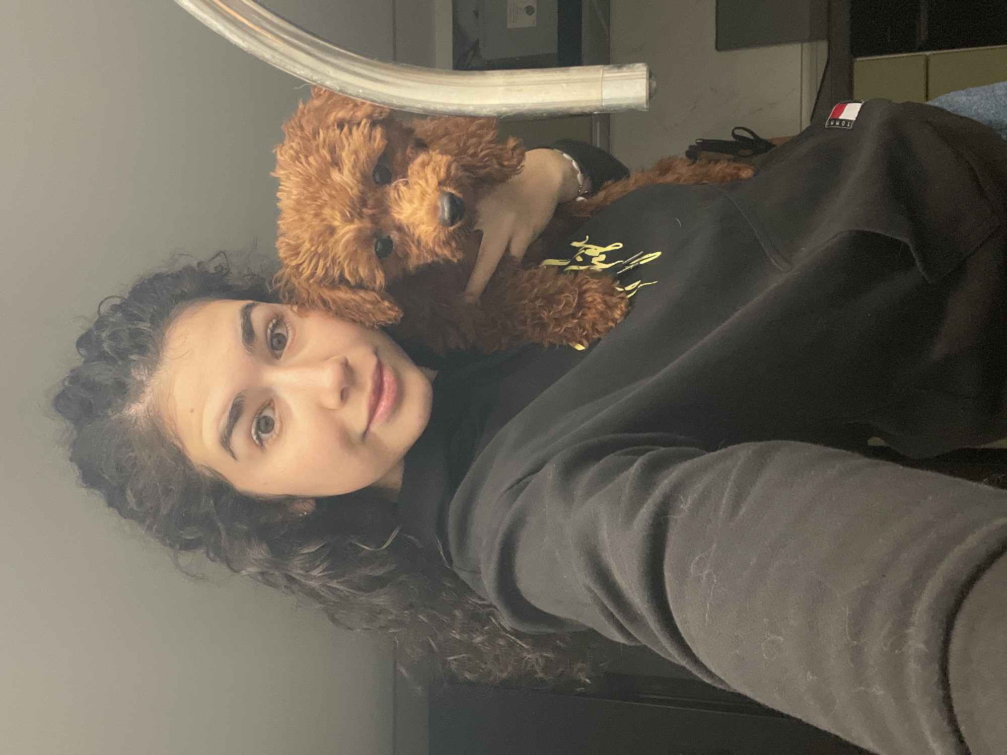 A selfie depicts a woman with curly dark hair, fair skin, and brown eyes. She is wearing a black hoodie with yellow lettering. Cradled on her chest is a small, brown poodle-like dog. A cylindrical metal object is visible above the dog. The background is indistinct but appears to be an indoor setting, possibly a bathroom or bedroom, with neutral-colored walls. The location cannot be determined to be Sofia, Bulgaria.