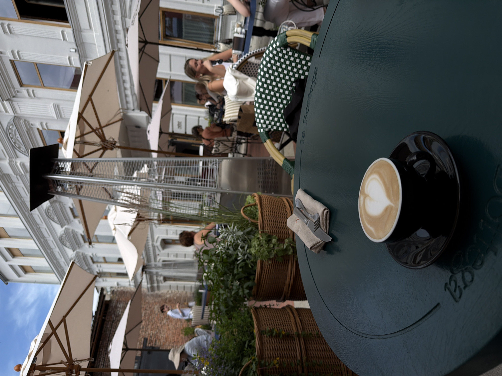 The media file captures an outdoor seating area of a cafe on a sunny day. The scene is dominated by a dark green circular table in the foreground, on which a black cup of coffee with latte art is placed. A napkin with cutlery is also on the table. In the background, several people are visible, some seated at tables, suggesting a relaxed atmosphere. Umbrellas provide shade over some of the seating areas. The architecture of the buildings in the background, with their windows and decorative elements, indicates an urban setting. The overall mood appears to be leisurely and social. The visible text on the table reads "La Bottega".
