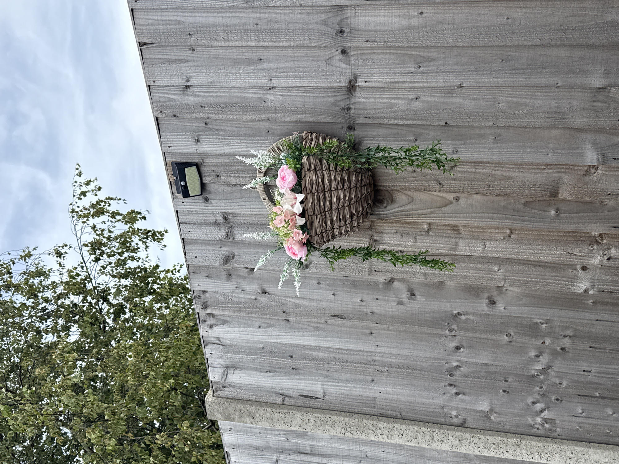 A weathered wooden fence forms the backdrop for a hanging basket filled with artificial flowers and trailing greenery. The basket, made of woven material, is attached to the fence with visible fixings. Above the fence, the branches of a tree laden with green leaves are visible against a cloudy sky. A solar-powered light fixture is mounted on the fence to the left of the basket. A concrete fence post is partially visible on the far left of the image. The scene appears to be in a garden or outdoor area, likely a residential setting in Great Yarmouth, United Kingdom.