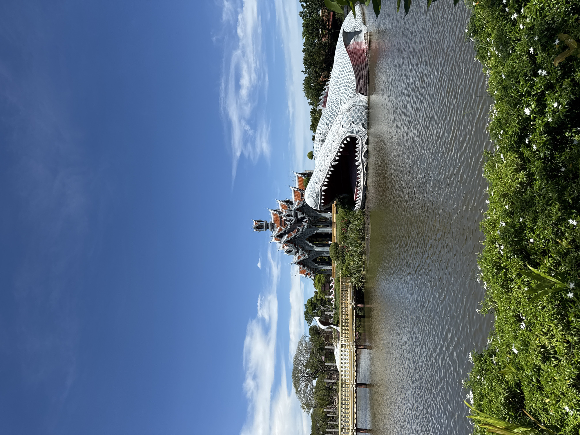 The image shows the Dragon Head Structure at Ancient City in Bang Pu Subdistrict Municipality, Thailand. The sculpture is in the water, facing to the right. To the left, across the water, a detailed building is visible. The sky is blue with scattered white clouds. Green vegetation is on the right side of the frame and a bridge is in the lower-left corner.