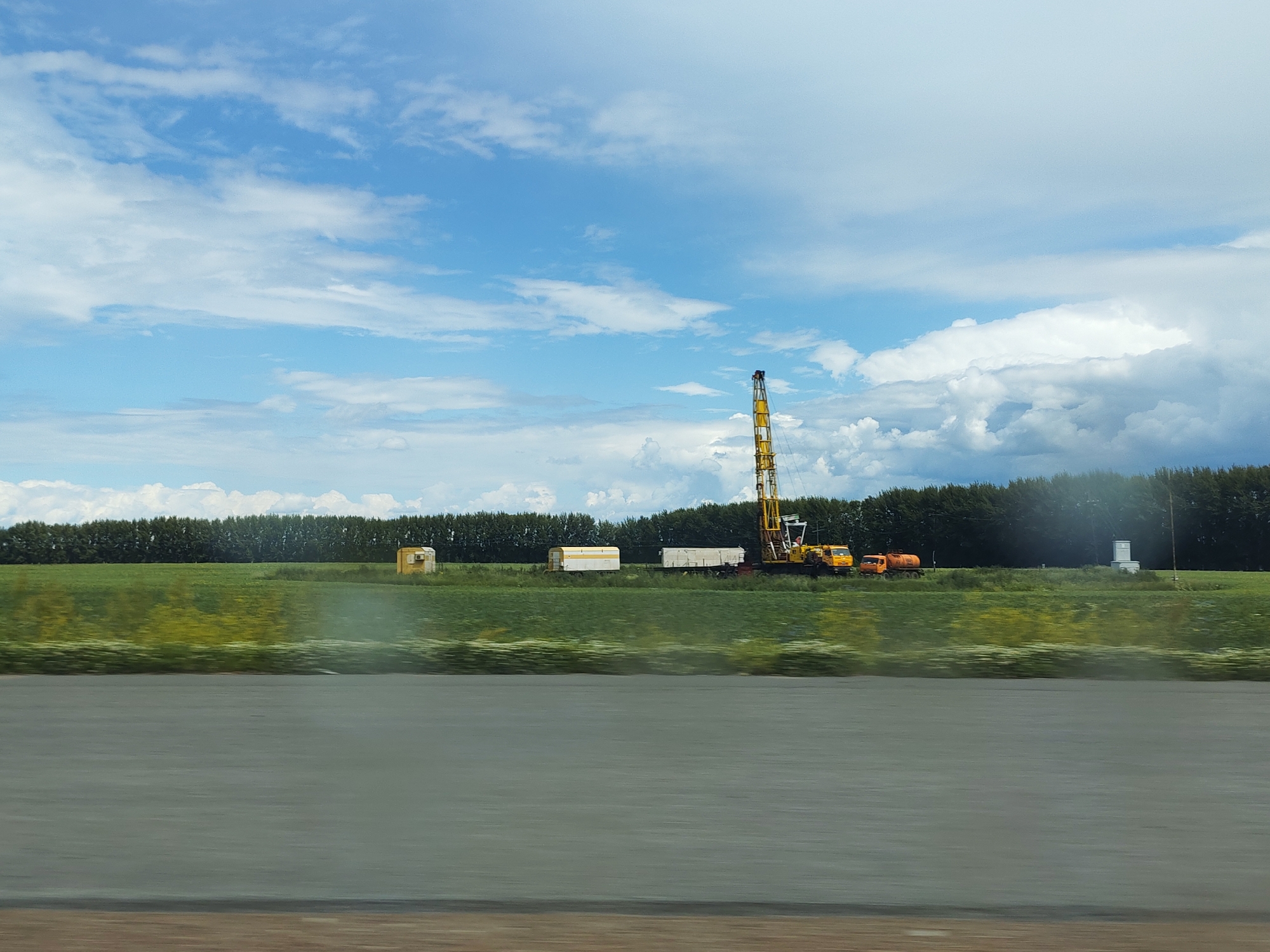 This image depicts a rural landscape on a cloudy day. The scene features a field of green grass with some yellow wildflowers, bordered by a dense line of dark green trees in the background. In the middle ground, several objects are present, including a tall yellow drilling rig, two trailers, and an orange truck. To the far left of the trailers, there is a small yellow building. The sky is a bright blue with scattered white clouds, some of which are large and puffy. The overall impression is of an industrial or construction site situated in a natural environment. The time of day appears to be daytime, likely late morning or afternoon, given the brightness of the sky. The weather seems to be fair, with a mix of sun and clouds. No people are visible in the image. The location context provided is Sarapul, Russia, which aligns with a landscape that could be found in that region. There is no visible text in the image.