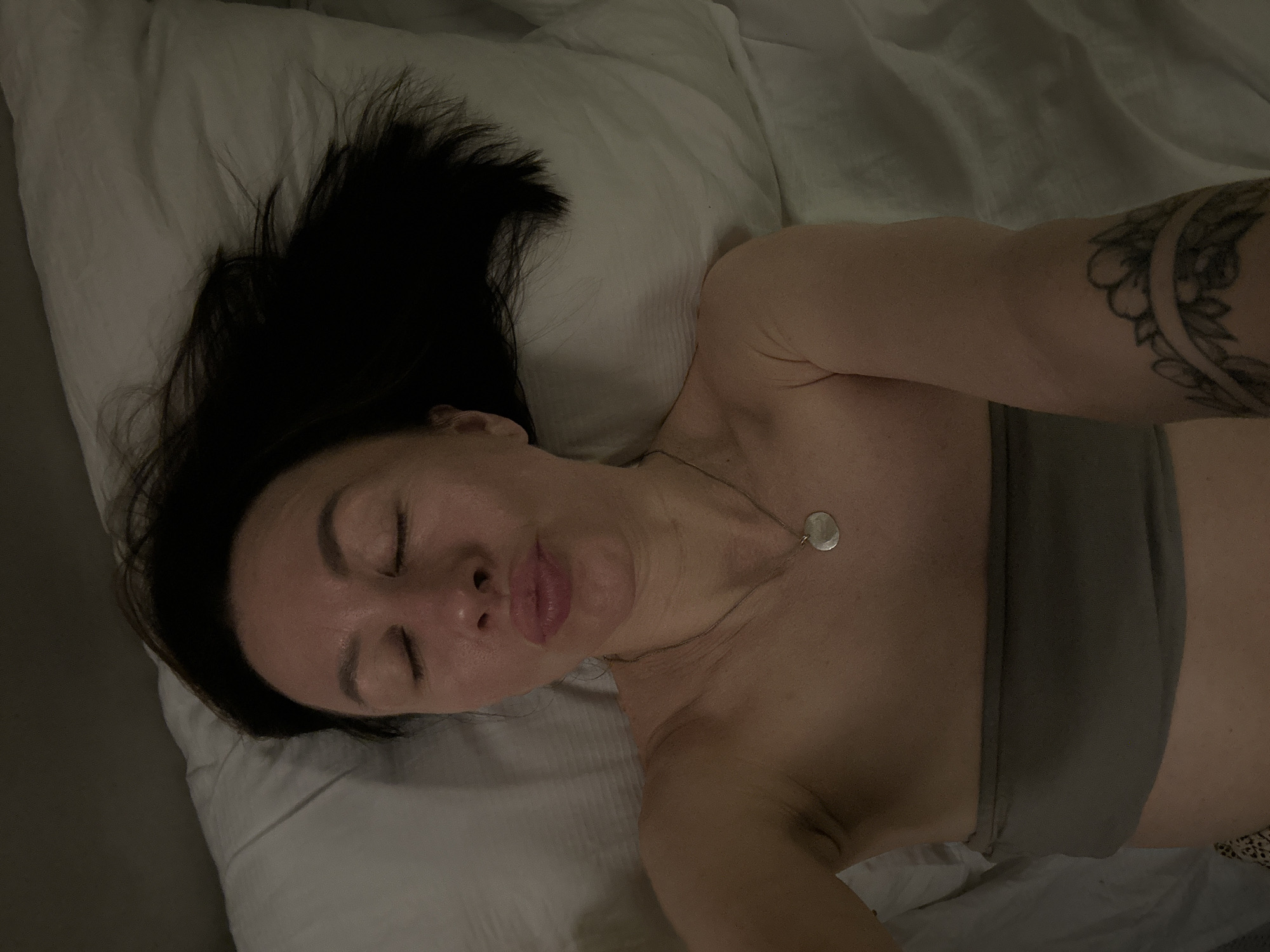 A woman with dark hair is lying on a white pillow and a white sheet. She is wearing a gray top and a silver necklace. Her eyes are closed, and her mouth is slightly pursed. Her left arm is visible, displaying a tattoo. The location in the city of Votkinsk, Russia is unknown.