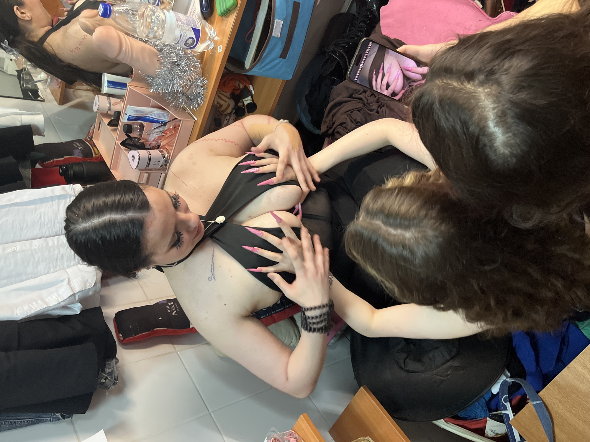 In this image, three individuals are gathered around a table laden with various items. One person, lying on their back, is wearing a black outfit and has long, sharp fingernails. They are holding their hands over their chest, with another person's hands also visible, touching their body. A third person is partially visible in the foreground, their hair obscuring their face.
The table is cluttered with personal belongings, including a bottle of water, a pink organizer filled with cosmetics, and what appears to be a sex toy. A silver tinsel garland is also present. The background shows shelves stocked with folded clothes and bags, suggesting a personal space like a bedroom or dressing area. The lighting suggests it is daytime. The overall mood is intimate and potentially suggestive, given the items and poses. There is some visible text on a product in the bottom right of the image, but it is too blurry to read clearly. The location is identified as Kazan, Russia.