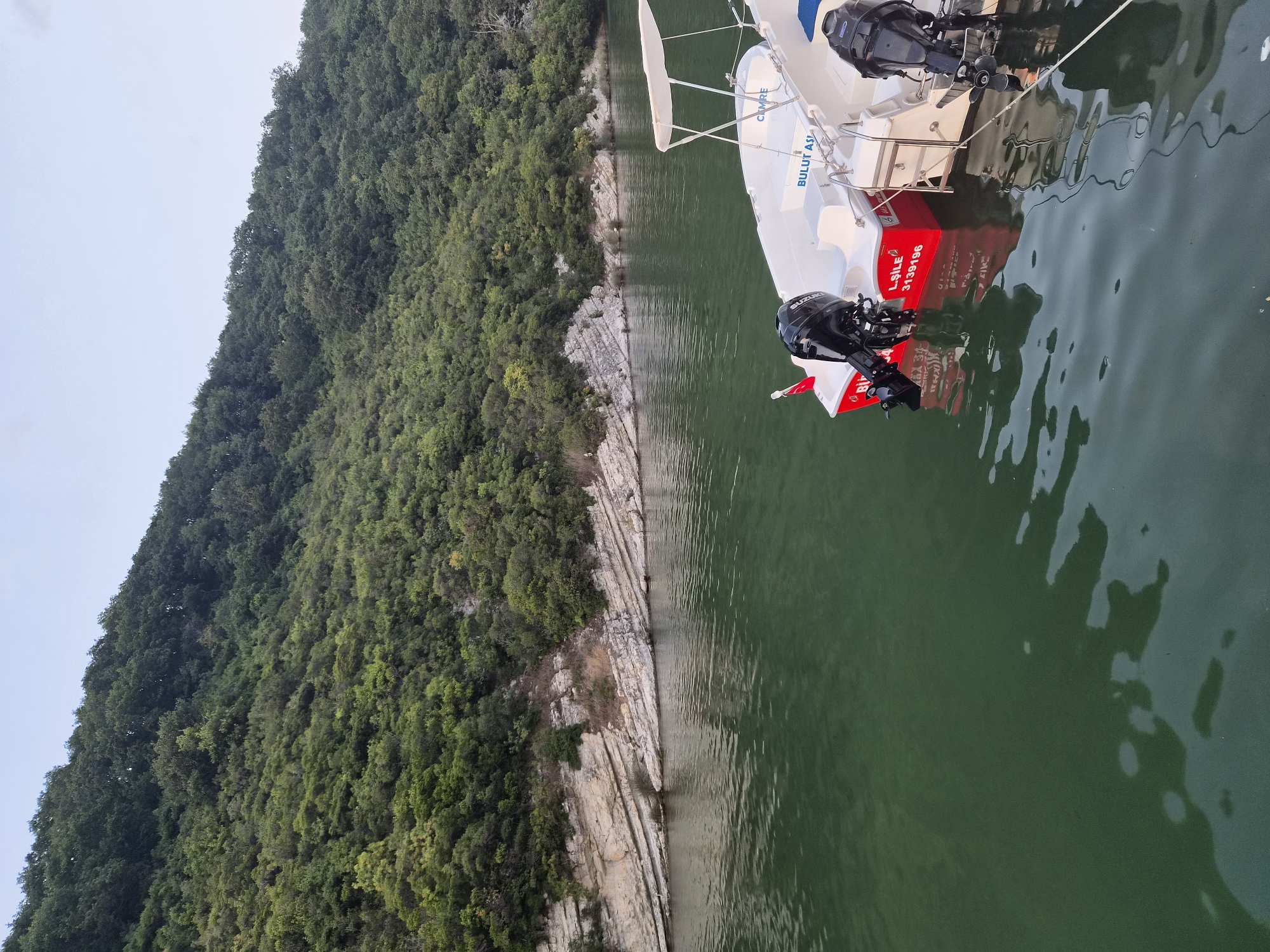 This is a photograph taken in Şile, Turkey, showing a scenic view of a calm, green body of water leading to a forested hillside. Two white boats with outboard motors are docked in the foreground. The boat on the left is red with white accents and has a visible serial number. The boat on the right has a canopy and is also white. The hillside is covered in dense, green foliage, with visible rock formations along the waterline. The sky is a pale blue with some cloud cover. The overall impression is one of natural tranquility.