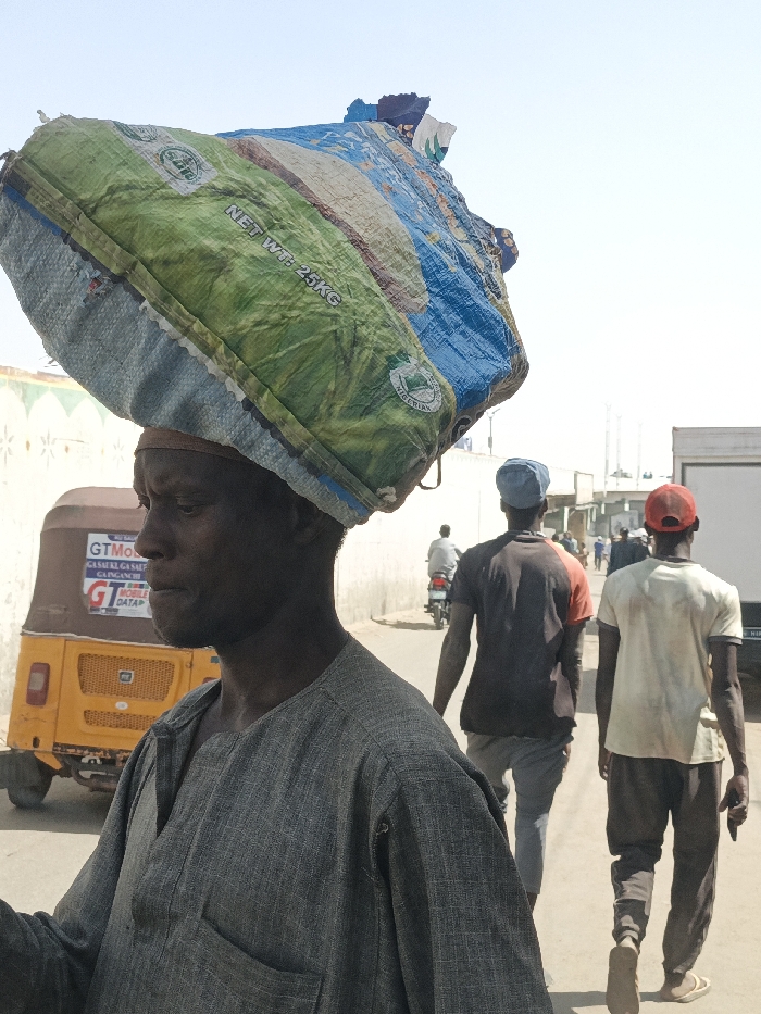 The scene depicts a daylight street view in Fagge C, Nigeria. In the foreground, an adult male, with dark skin and a neutral expression, is seen in left profile. He wears a grey long-sleeved garment with a small chest pocket and has a light-colored band around his forehead. He carries a large, irregularly shaped bundle on his head, consisting of a green and blue woven polypropylene sack printed with "NET WT: 25KG" and additional text including "NIGERIAN".
Behind the foreground subject and to his left, a yellow-orange auto-rickshaw (tricycle) is parked. Its side bears commercial lettering, including "GTMobile", "GAS/KER/GAS", "SUPER", "GAGAICHI", and "GT DATAP".
Further down the unpaved street, three male figures are walking away from the camera. The closest wears a dark t-shirt with orange accents and a blue cap. Beside him, another wears a light-colored t-shirt, dark pants, and a red cap. Further beyond them, a male is seen riding a motorcycle. The background features light-colored walls, indicating buildings, and extends into a bright, clear sky. A white commercial vehicle, possibly a truck or van, is partially visible on the far right in the distant background.