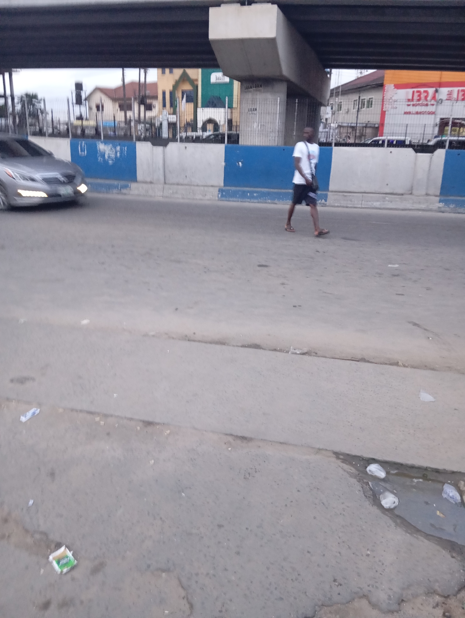 The image shows a street scene in Port Harcourt, Nigeria, possibly under an overpass. A silver car is driving on the left side of the road. A man is walking across the road from right to left. He is wearing a white t-shirt, dark shorts, and sandals, and carrying a bag slung over his shoulder. In the background, there are buildings and a large concrete structure that appears to be part of an elevated road or bridge. The road surface is asphalt and appears worn, with some litter visible, including a crumpled paper packet and some plastic wrappers. The sky is overcast.